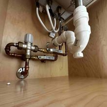 Under-sink view showing white plastic drainage piping and copper water supply lines with valves attached to wood panels.