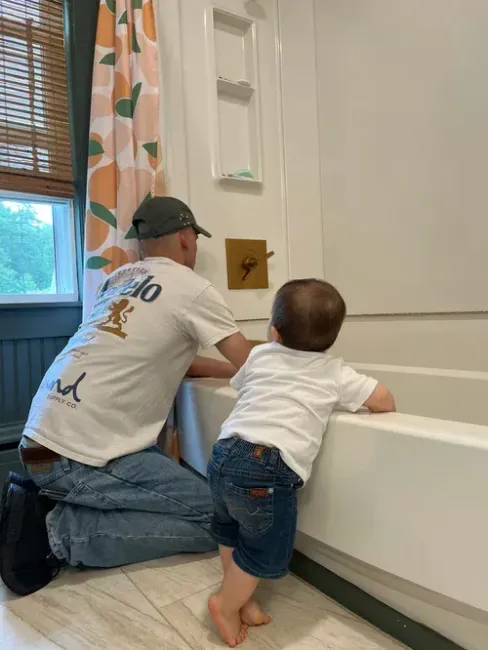 A person and a toddler kneel by a white bathtub, looking at a golden square mounted on the wall inside a bathroom.