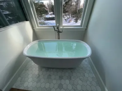 A freestanding white oval bathtub filled with water sits on a light gray hexagonal tiled floor in front of a window.