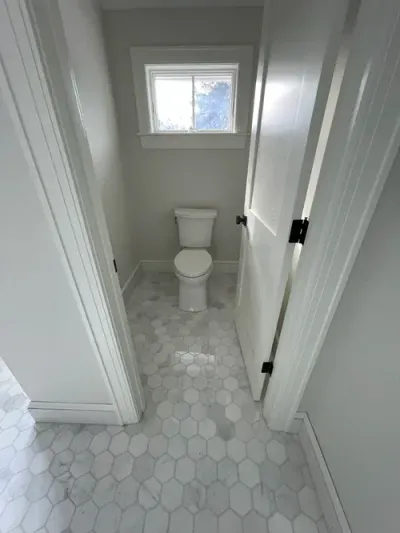 A small, neutral-toned toilet room featuring light gray walls, white hex floor tiles, and a white toilet.