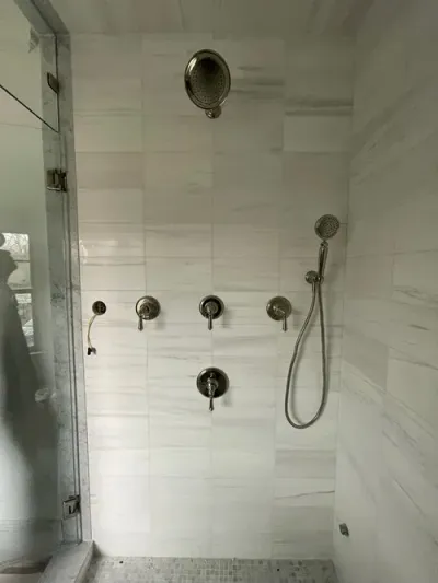 A modern shower stall with light marble-patterned tiles, a rainfall showerhead, a handheld sprayer, and wall-mounted controls.