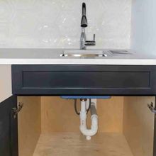 A close-up of a kitchen sink with a chrome faucet, dark gray cabinet door, and visible white P-trap plumbing underneath.