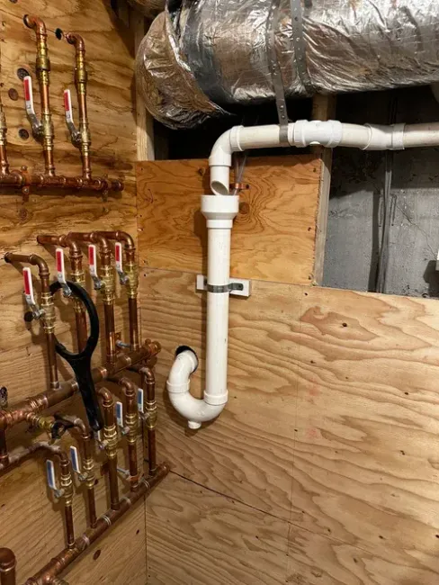 A plumbing setup with copper manifold valves on the left and a white PVC drain pipe with a trap attached to plywood walls.