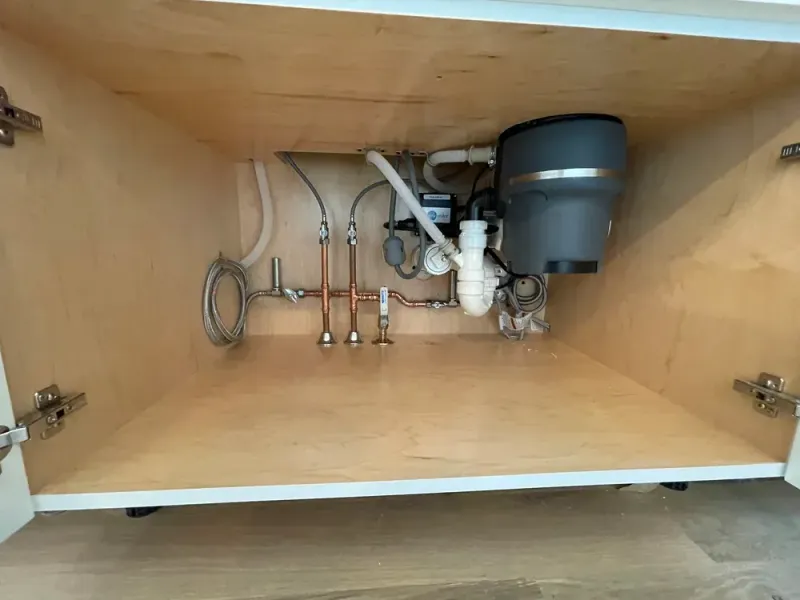 Under-sink cabinet with a garbage disposal, plumbing pipes, and water supply valves against a light wood interior.