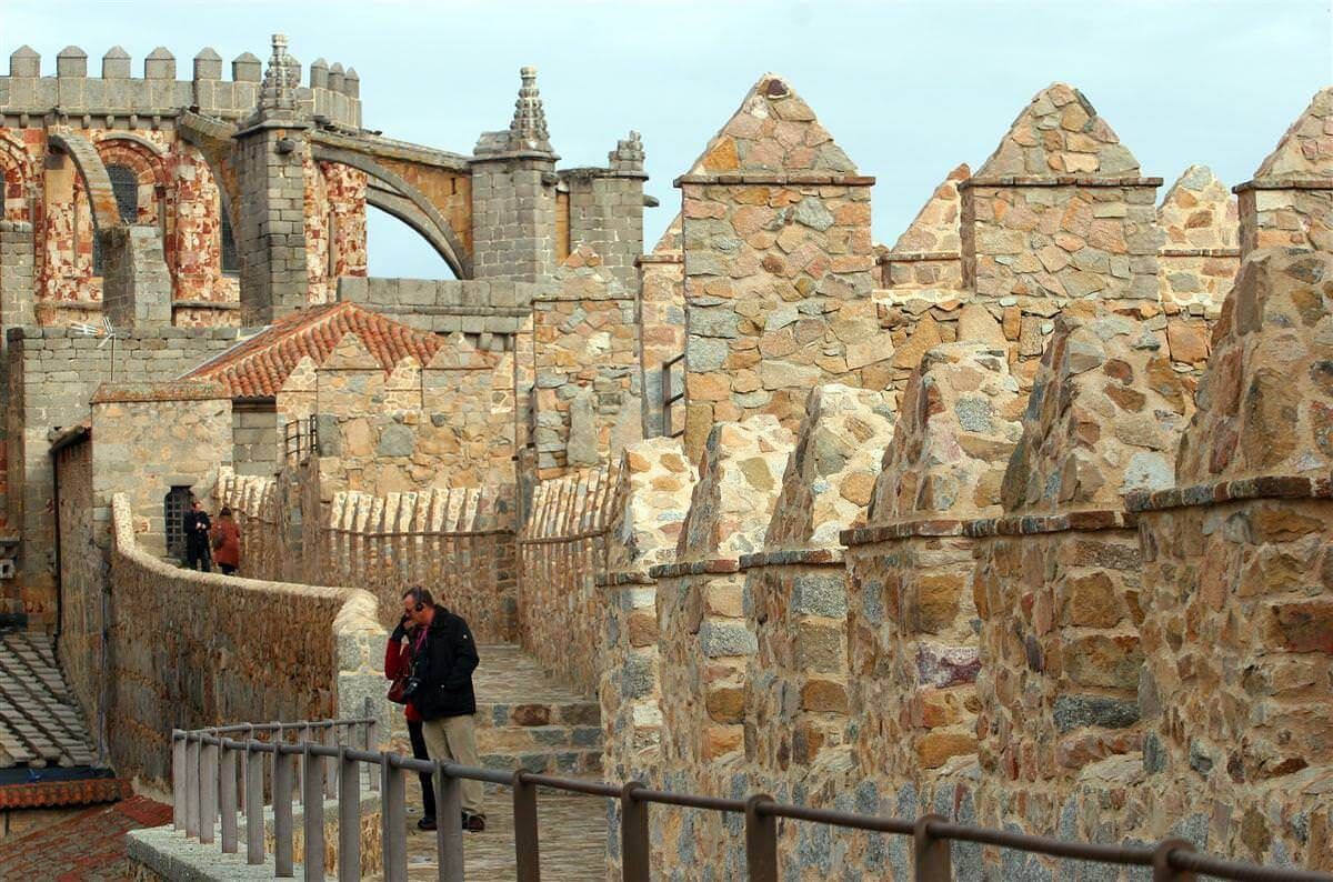 Murallas y almenas de piedra de una fortaleza, con un estrecho sendero por el que caminan algunas personas bajo un cielo azul.