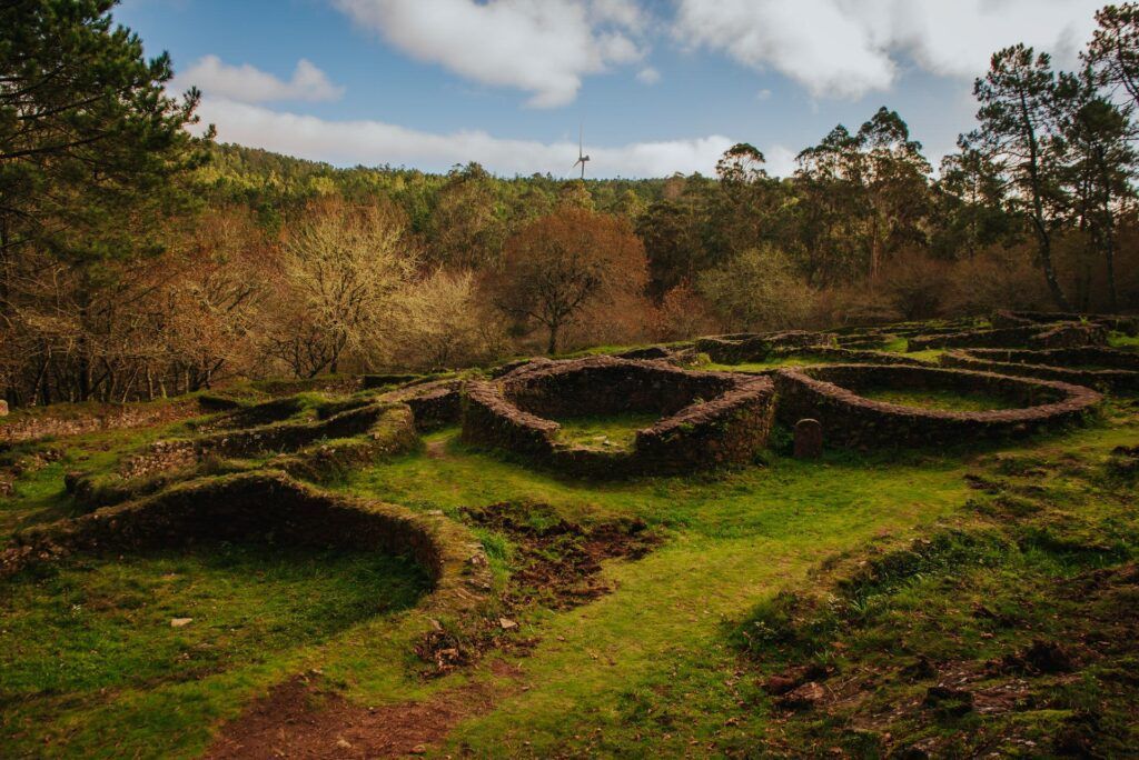 Ruinas cubiertas de hierba de muros bajos de piedra en un paisaje boscoso bajo un cielo parcialmente nublado.