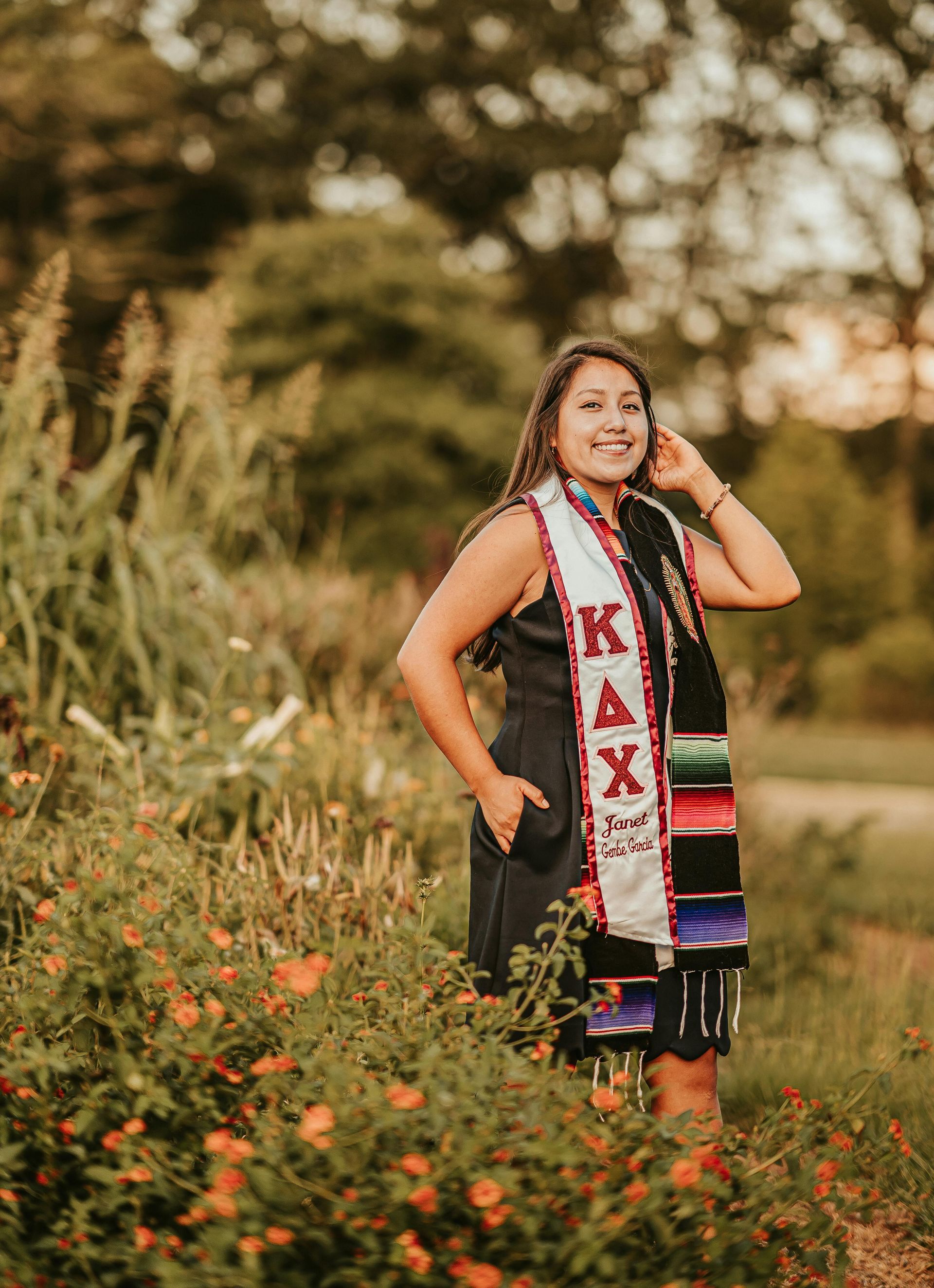 Woman in graduation sash smiling outdoors, surrounded by plants.