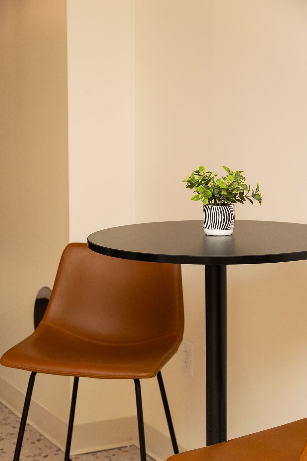 A brown chair sits next to a small, round black table topped with a potted plant against a neutral wall.