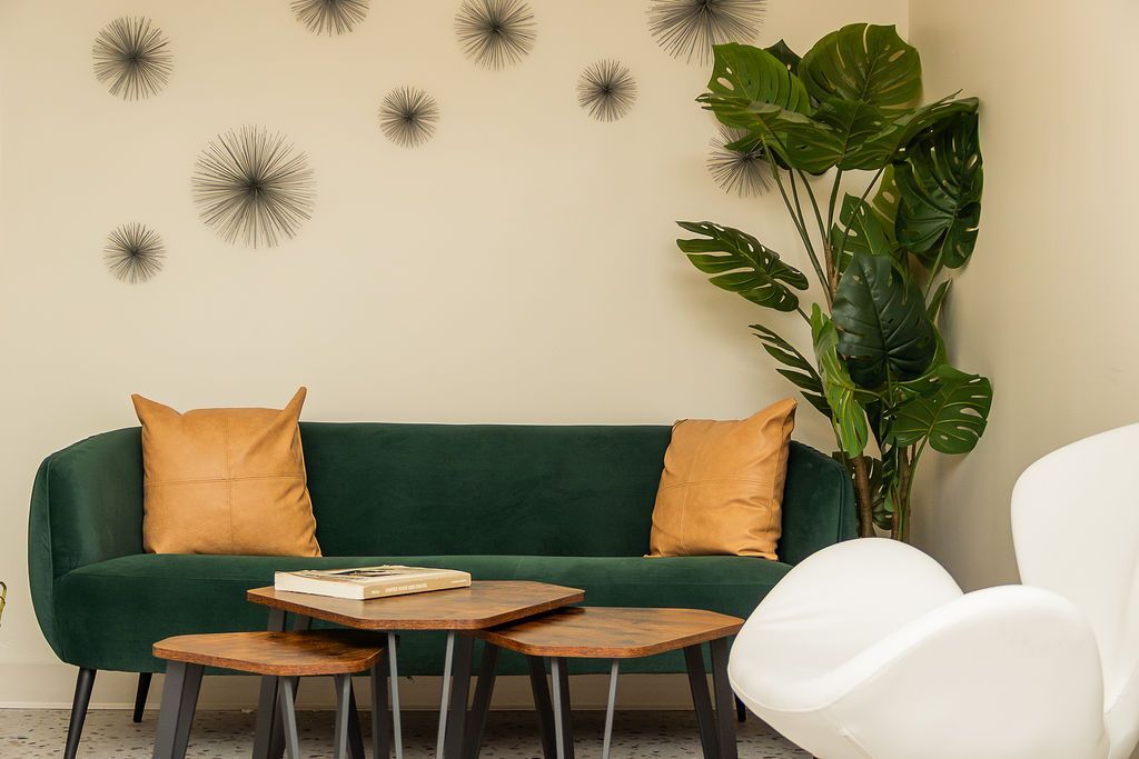 A green velvet sofa with tan cushions, wooden coffee tables, and a large monstera plant in a corner with wall decor.