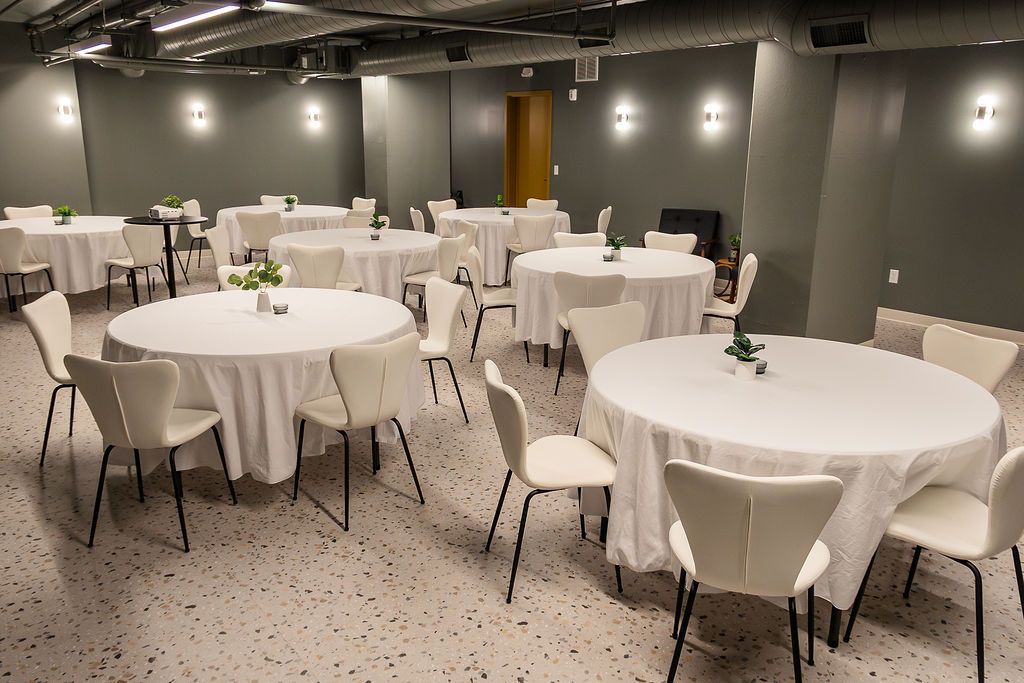 A spacious room set up for a formal event with multiple round tables covered in white cloths and modern white chairs.