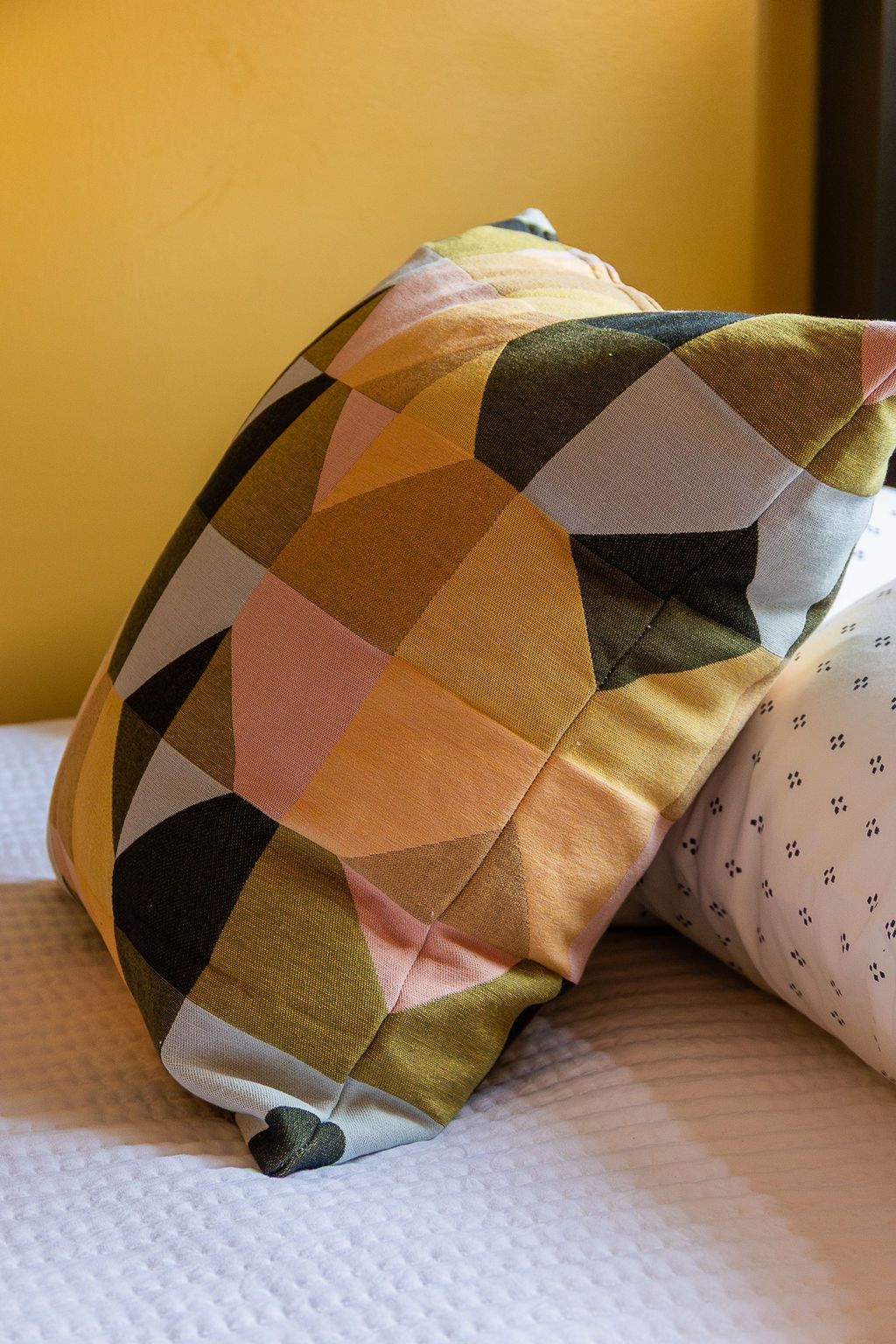 A decorative throw pillow with a geometric pattern in olive green, pink, and yellow, sitting on a white bed.