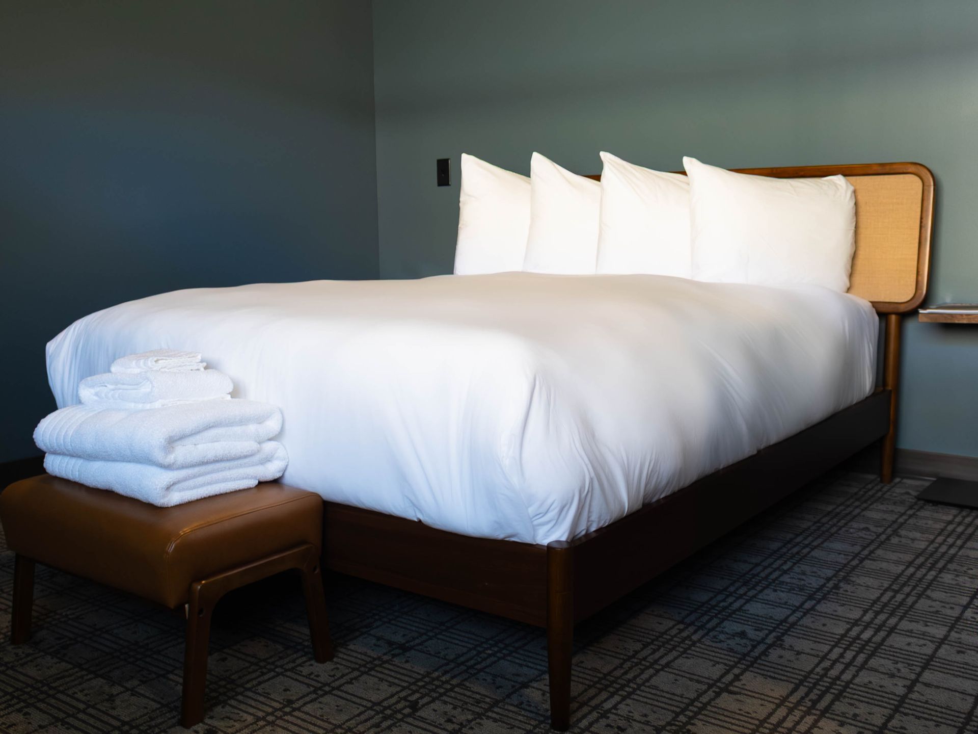 A hotel room with a neatly made bed featuring four white pillows, a stack of white towels, and a brown bench at its foot.