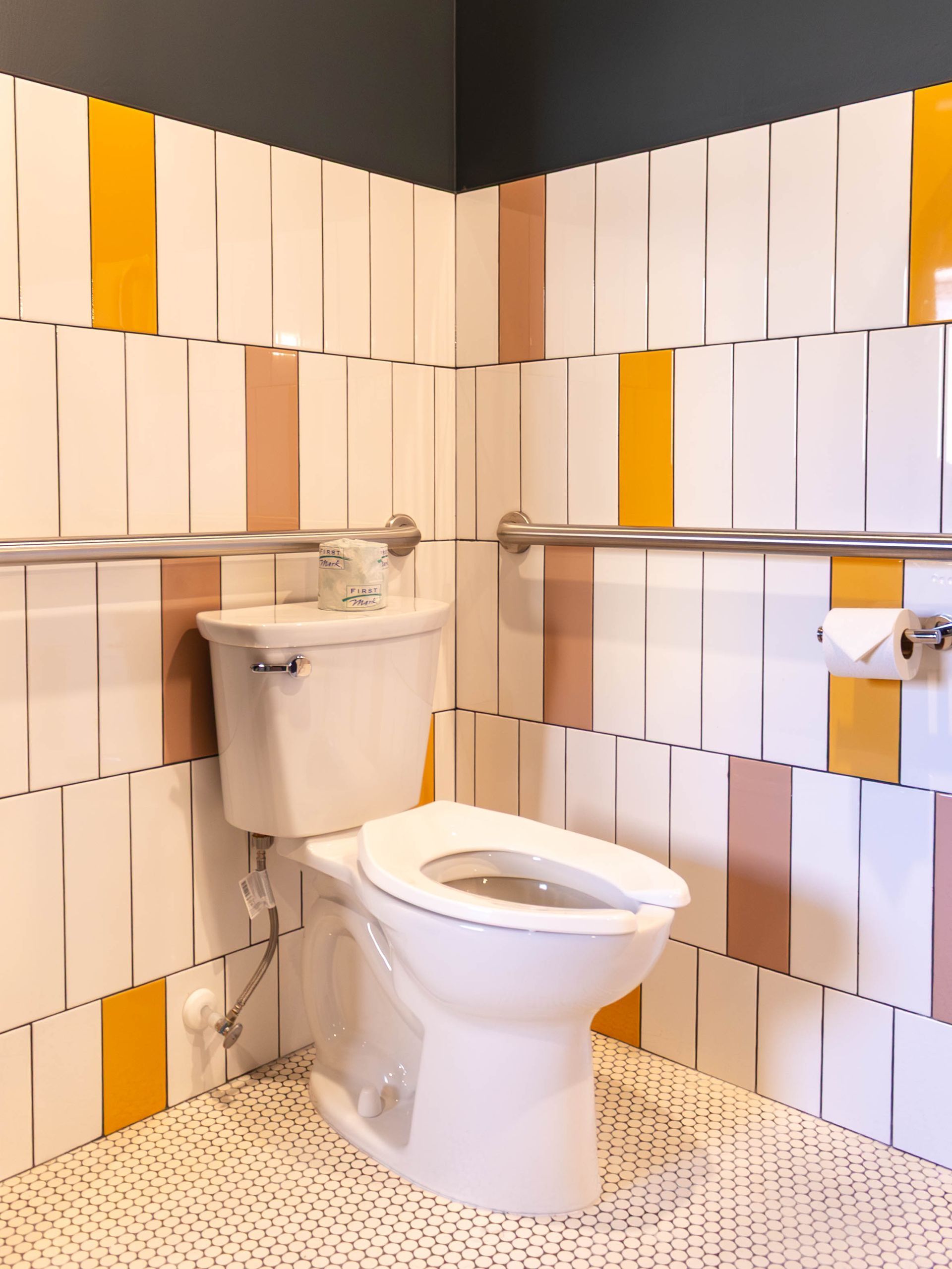 A toilet in a bathroom with patterned white, yellow, and tan rectangular wall tiles, a grab bar, and a toilet paper roll.