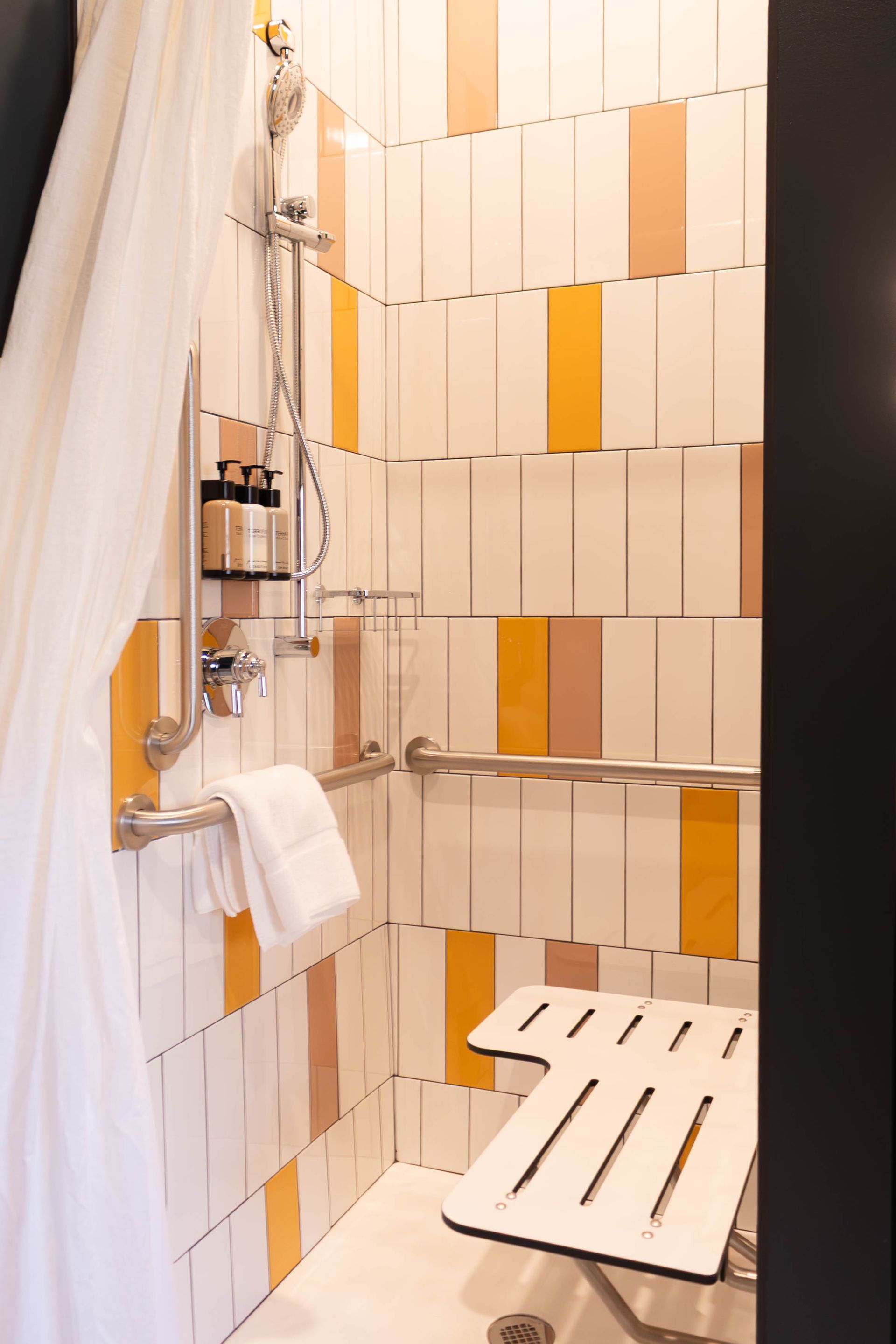 Accessible shower with a white fold-down seat, safety grab bars, a handheld showerhead, and yellow and white tiled walls.