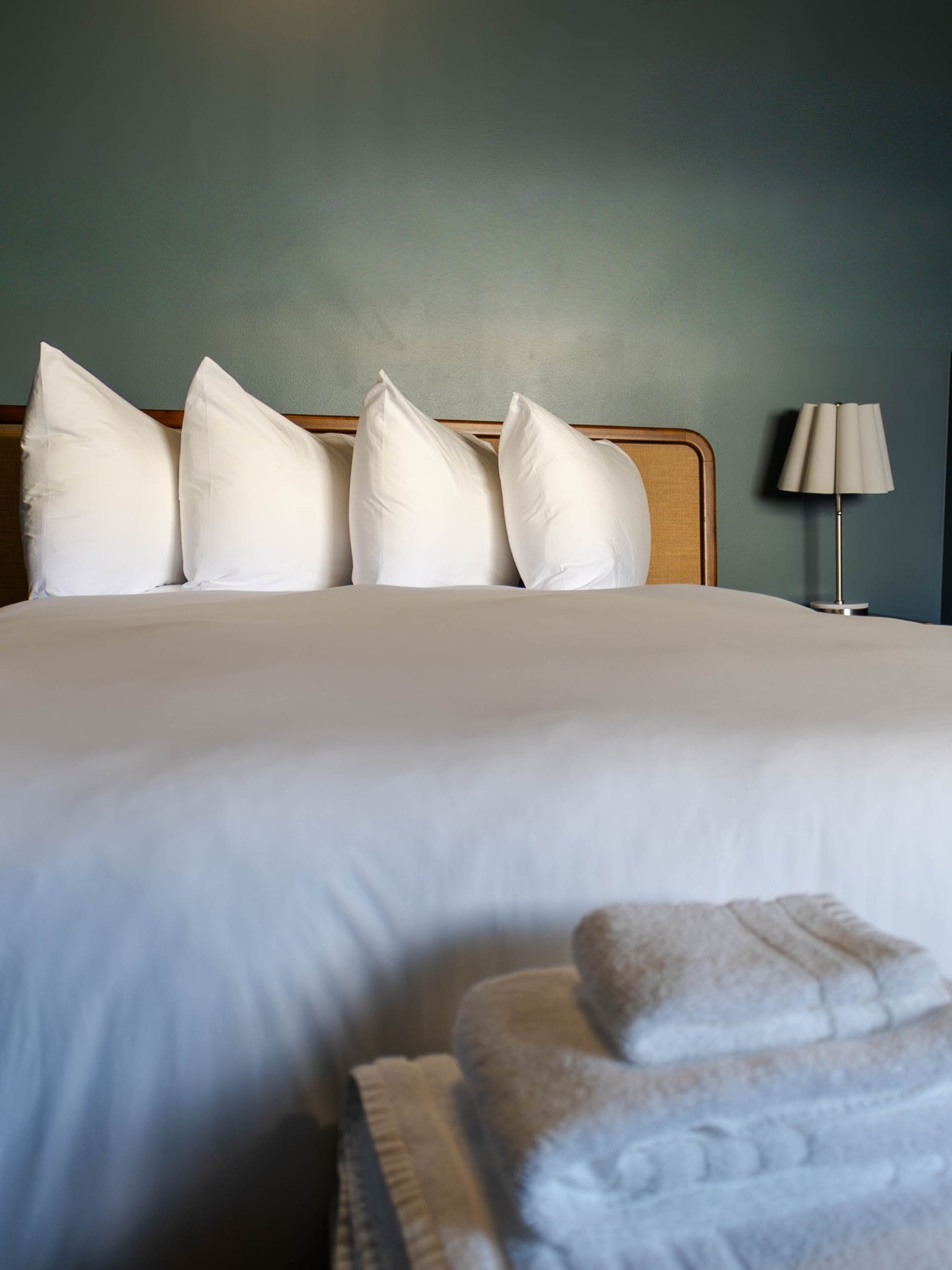 A white-made hotel bed with four fluffy pillows against a blue wall and a stack of folded white towels in the foreground.