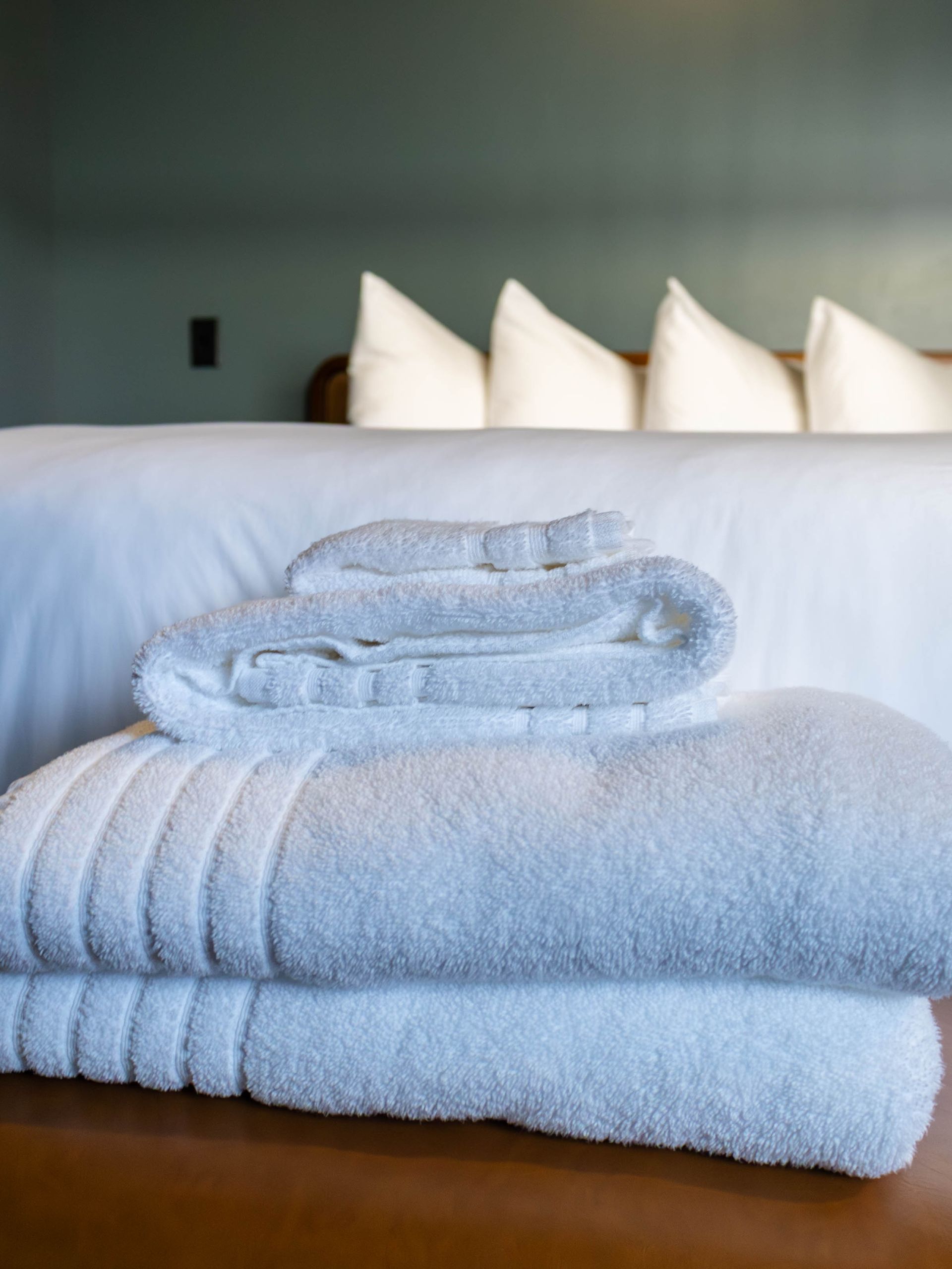 A stack of clean, white towels sits on a brown surface in front of a neatly made bed with four white pillows.