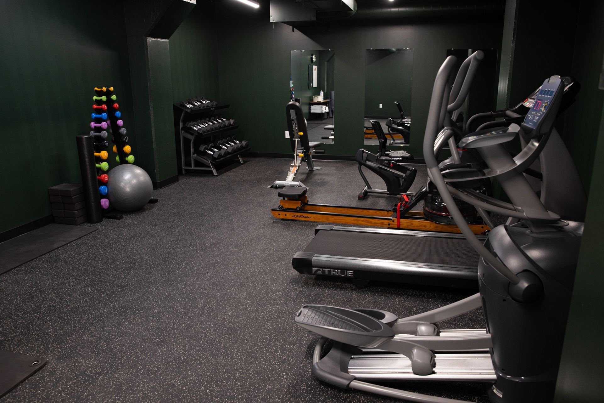 A gym with dark green walls, a black speckled floor, weight racks, an exercise ball, a rowing machine, and ellipticals.