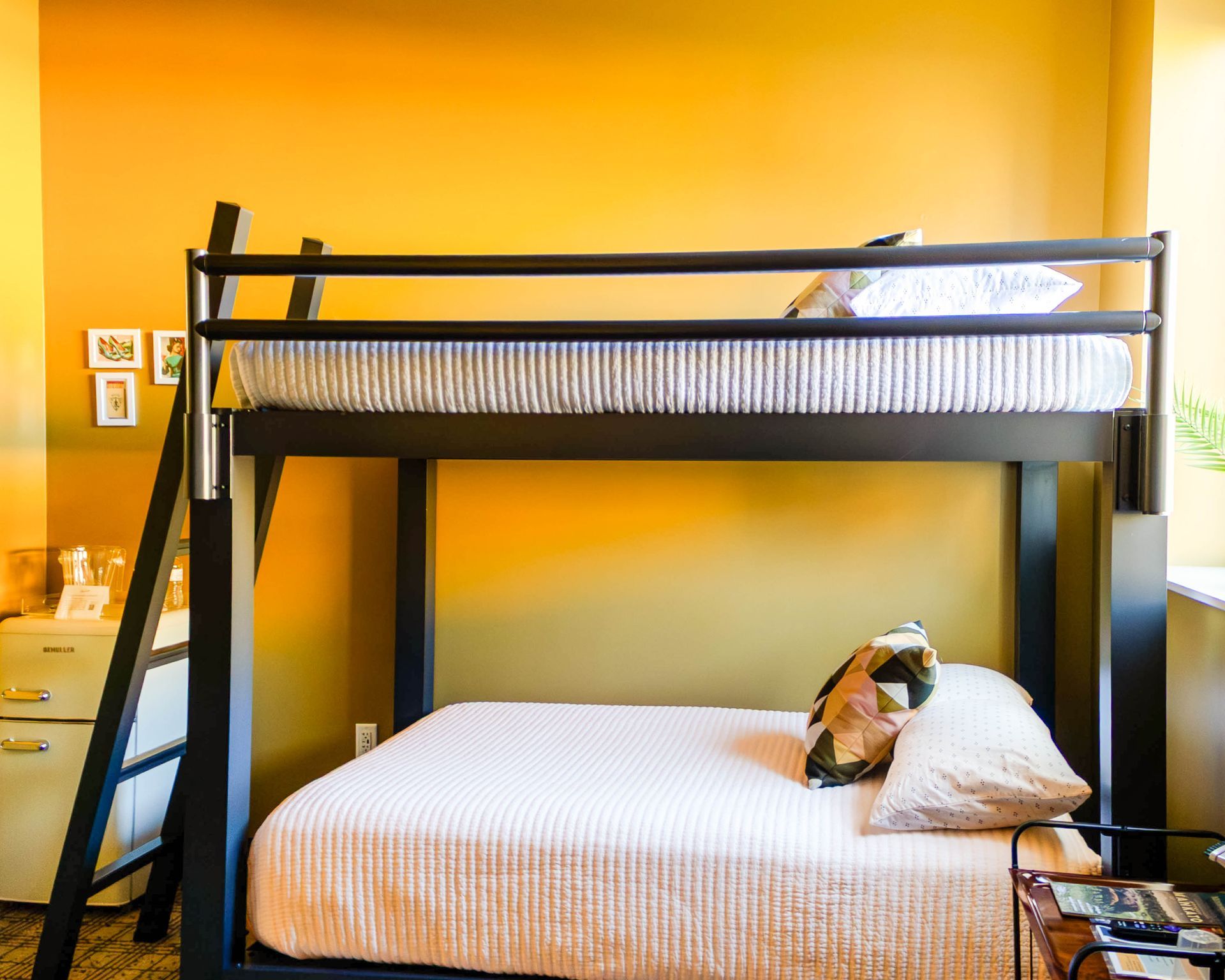 A hotel room with yellow walls, a black metal bunk bed, and a wall-mounted television above a small white mini-fridge.