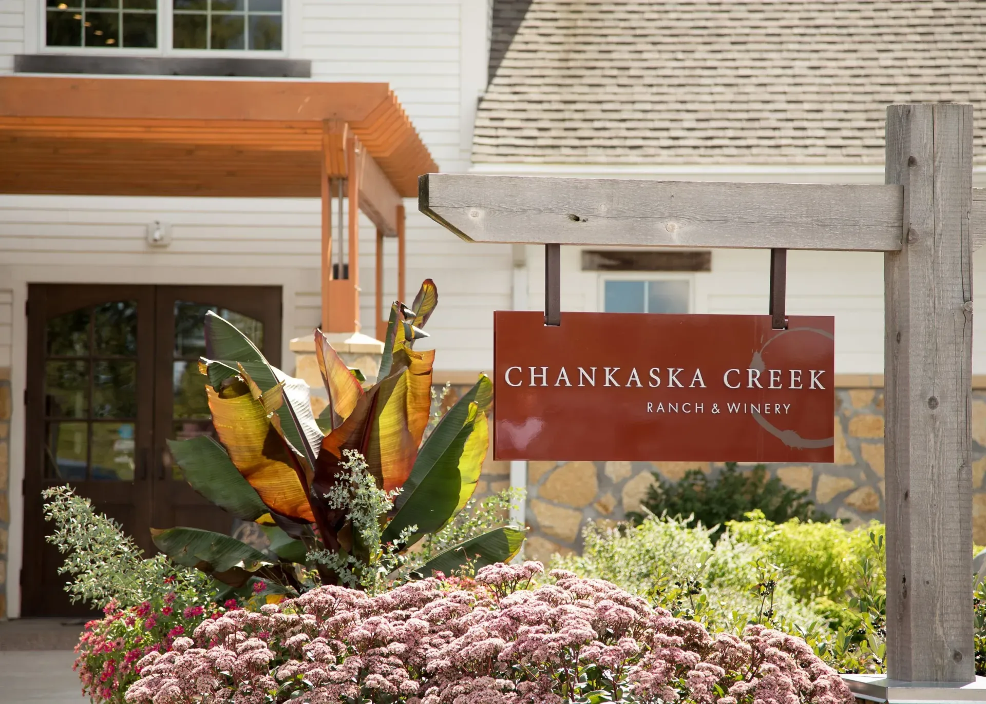 Chankaska Creek Winery sign on a wooden post in front of a white building with landscaping and flowers.