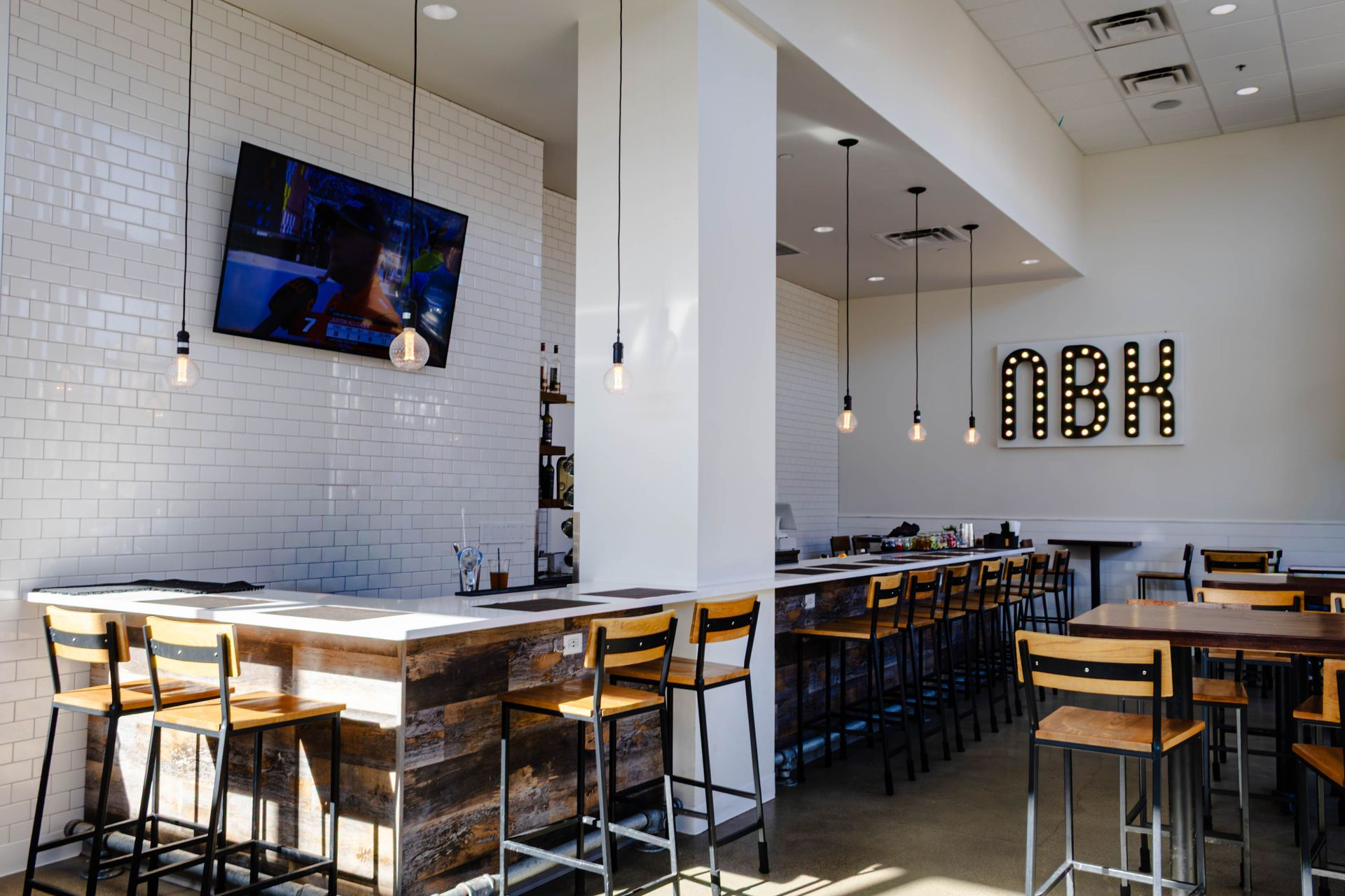 A modern restaurant bar with white brick walls, wooden bar counters, and high stools under pendant lights.