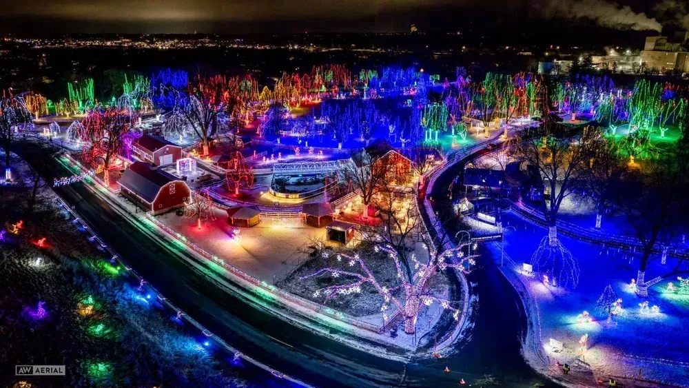 An aerial night view of a park filled with vibrant, multi-colored holiday light displays, paths, and a lit-up red barn.