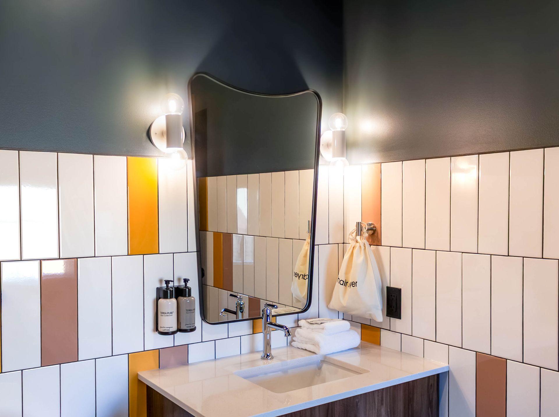 A bathroom vanity with a uniquely shaped mirror, white and colored tiles, wall-mounted soap bottles, and a hairdryer.