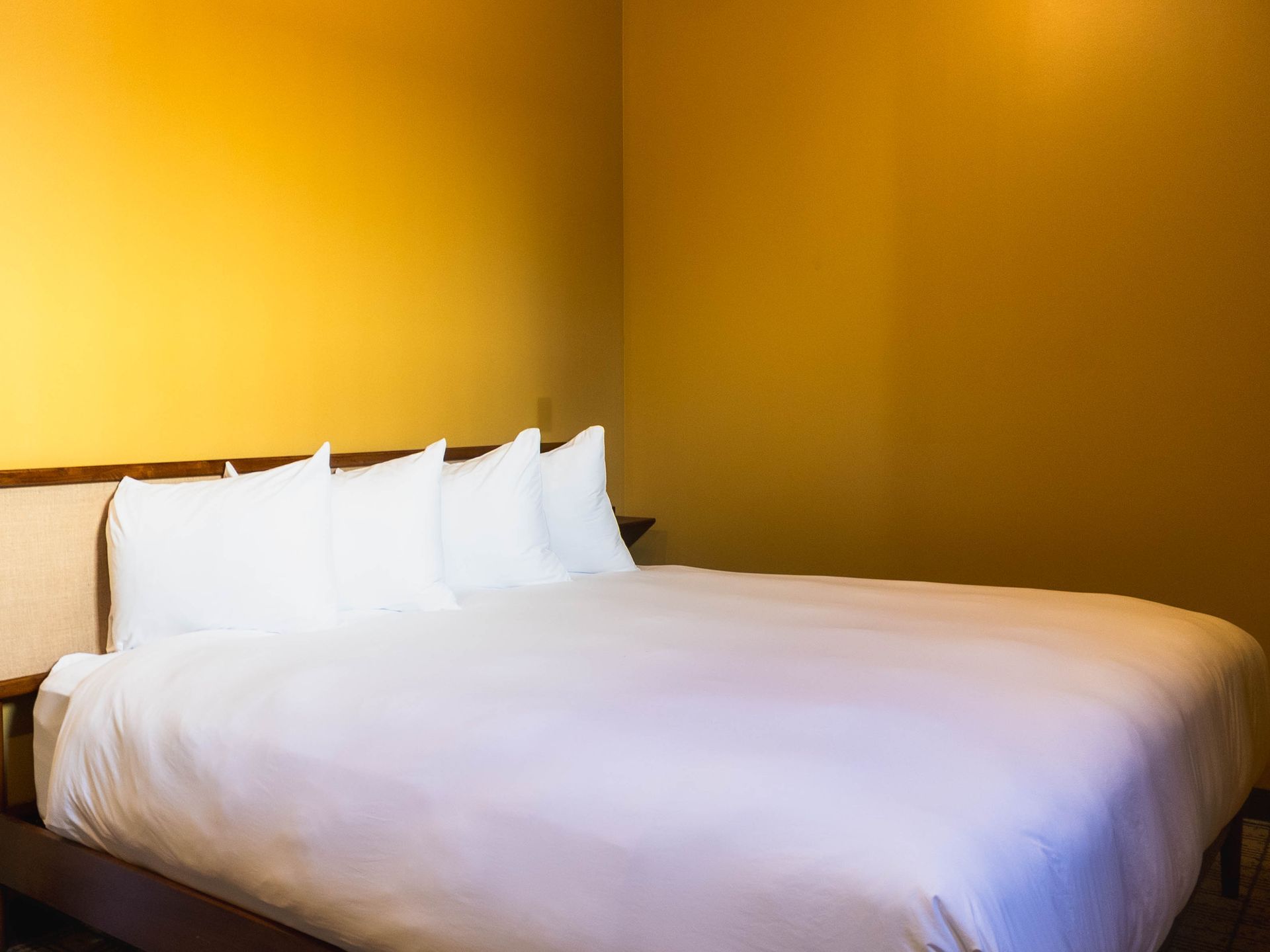 A neatly made bed with white pillows and bedding against plain, mustard-yellow walls in a simple bedroom.