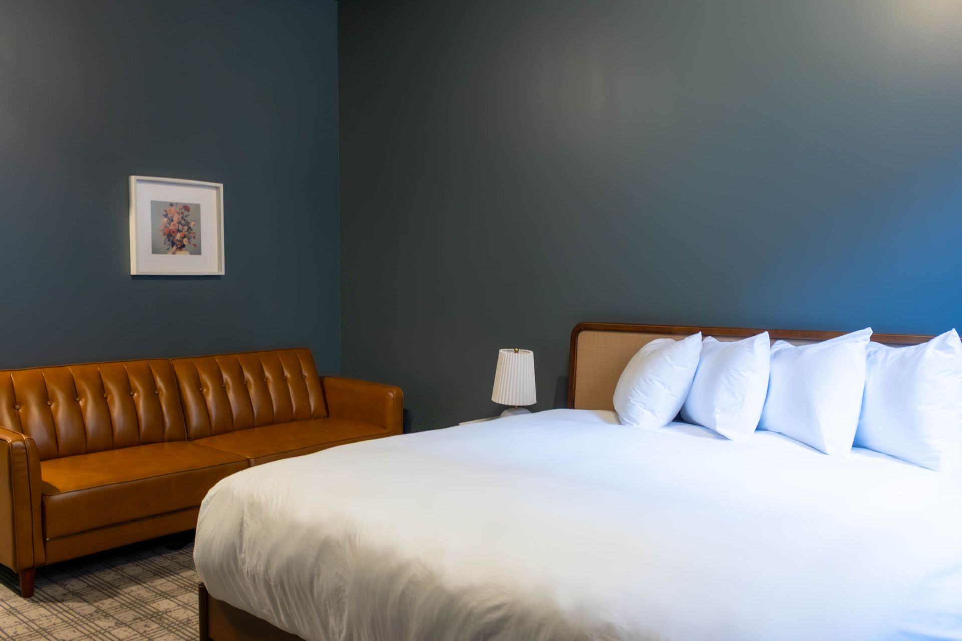 A bedroom with a tan leather sofa, a bed with white linens and pillows, and a small framed print against a dark blue wall.