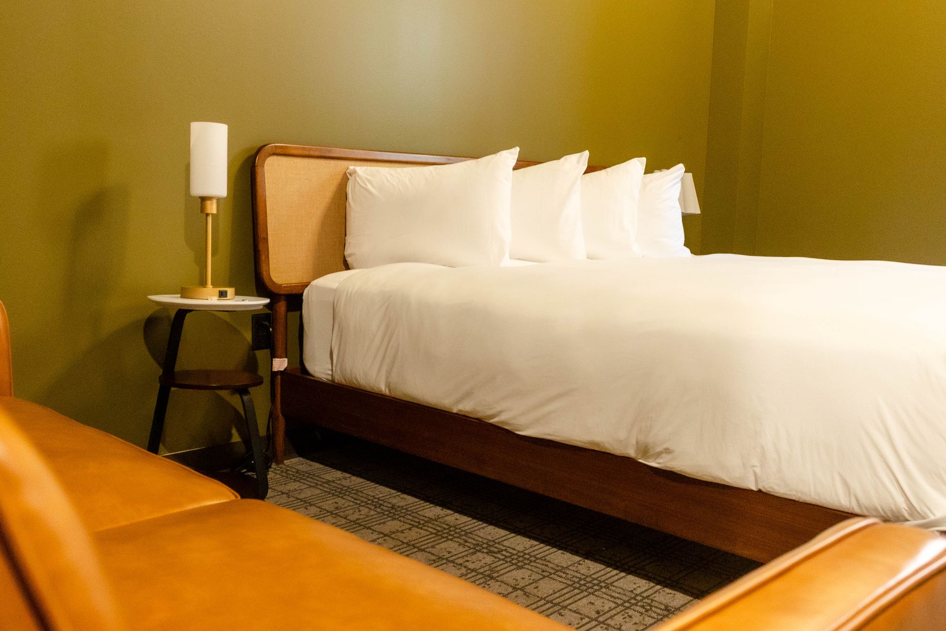 A view of a modern bedroom with olive walls, a bed with white linens, a wooden nightstand, a lamp, and a leather sofa.