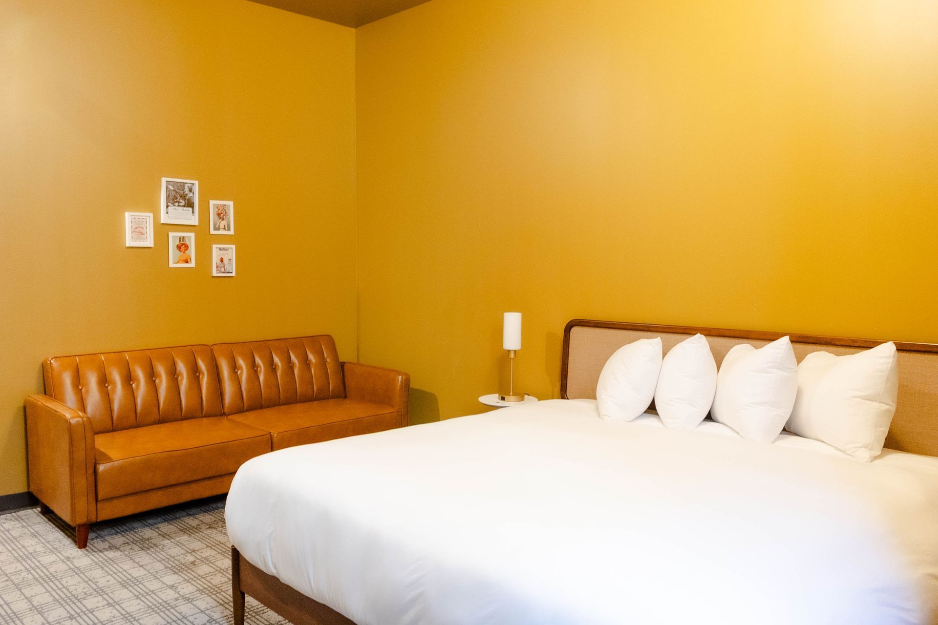A room with a white-bedded bed, a tan leather sofa, and small framed wall art against mustard-yellow walls.