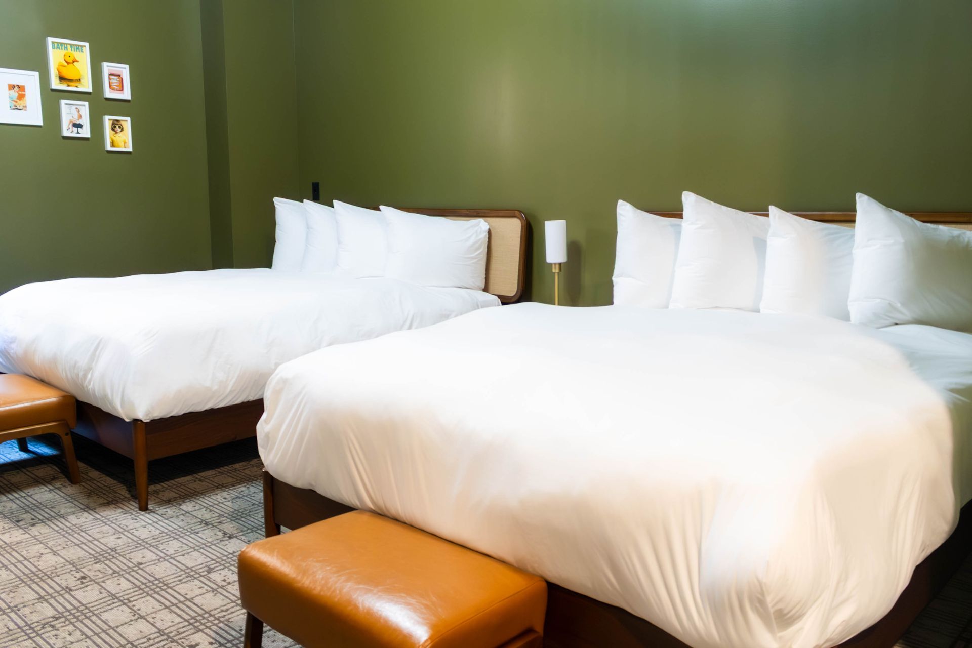 Two queen beds with white bedding and tan benches in a hotel room with olive green walls and small framed wall art.