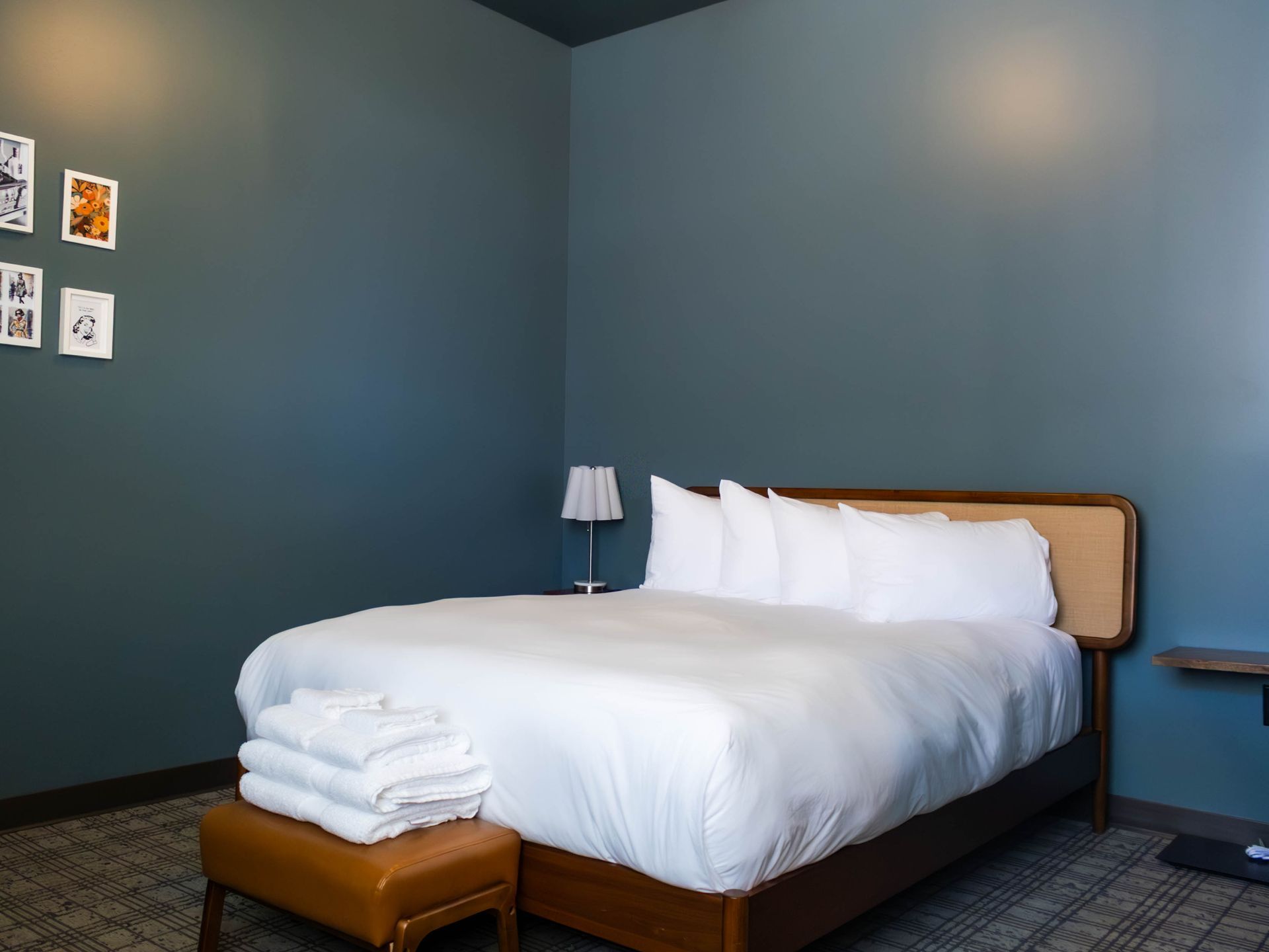 A modern hotel room with a neatly made bed, white linens, a tan footstool, and dark blue walls.