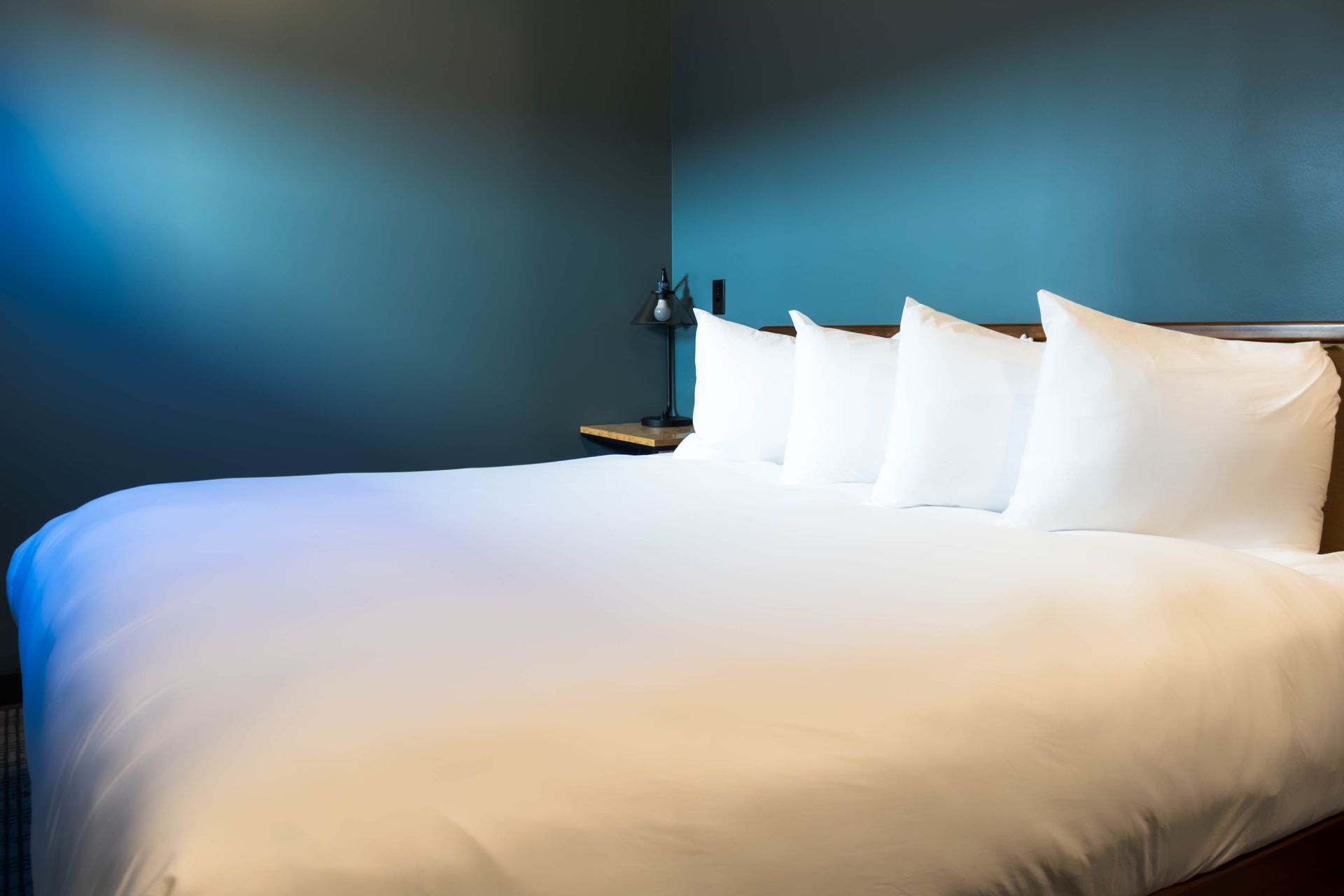 A made bed with four white pillows against a solid, dark teal blue wall in a minimalist hotel room.