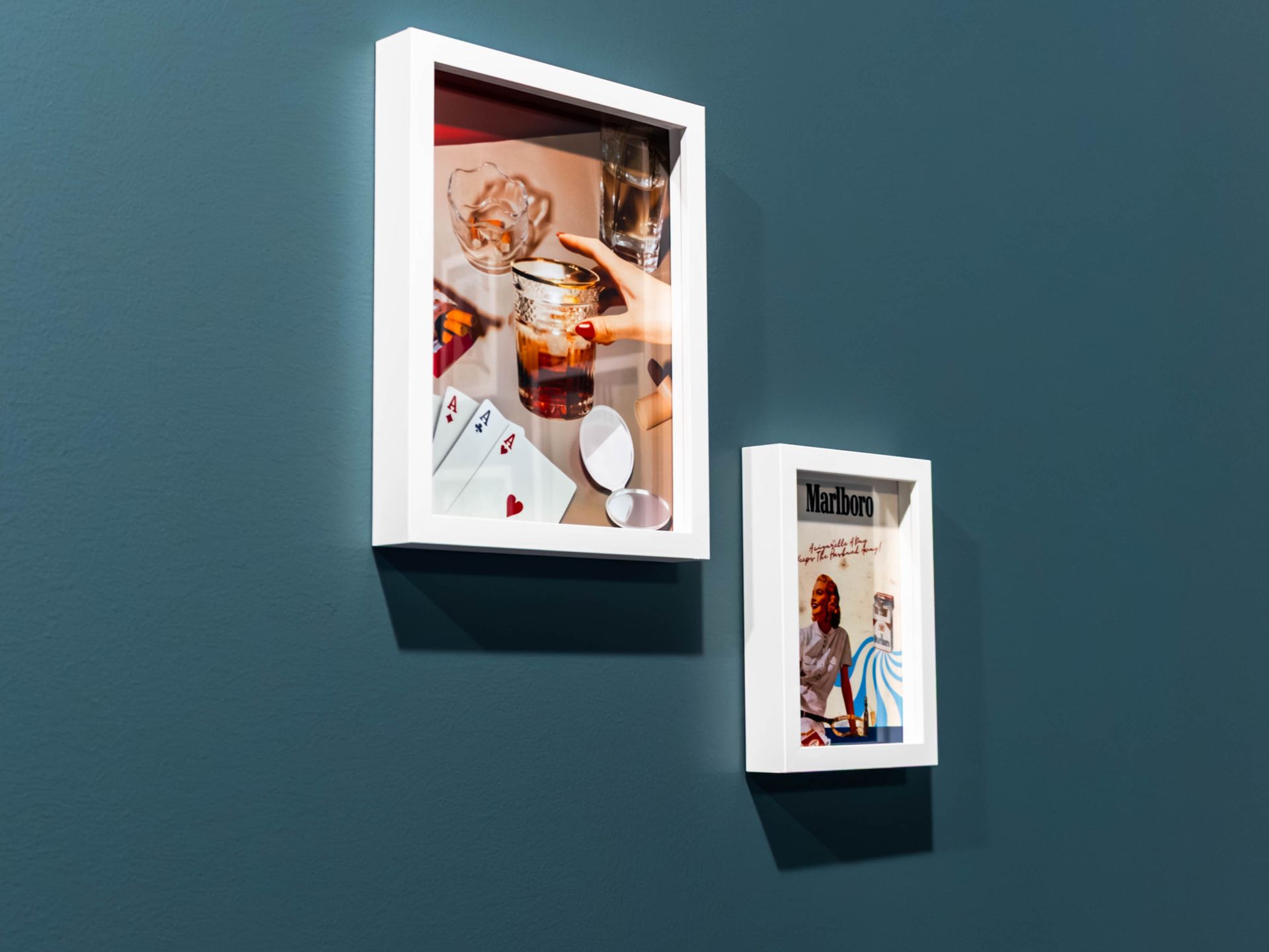Two white-framed shadow boxes hanging on a dark blue wall: one shows cards and a drink, the other a vintage advertisement.