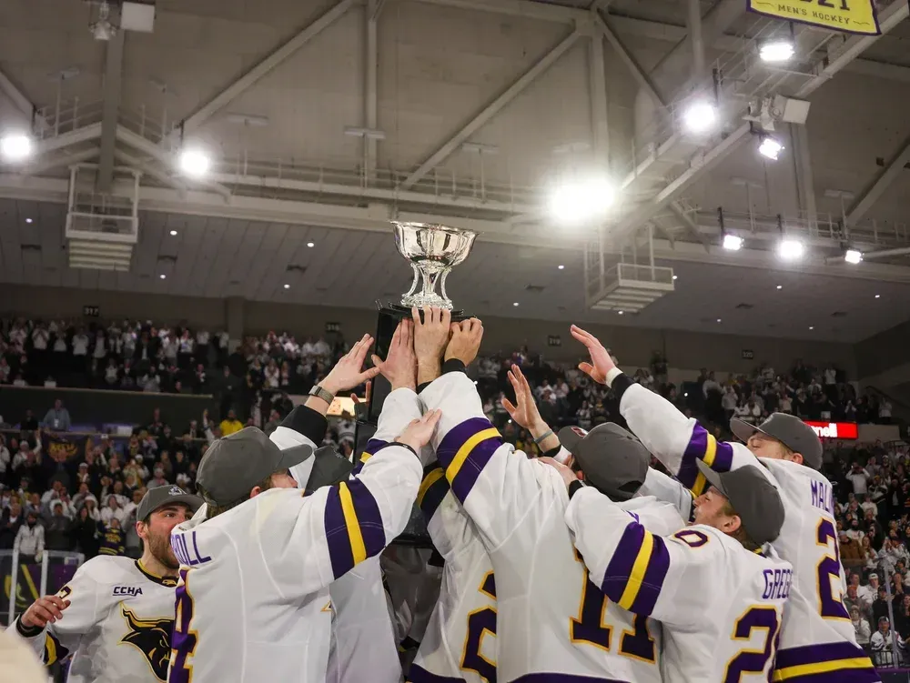 Hockey players in white and purple jerseys hoist a silver championship trophy in a crowded indoor arena.