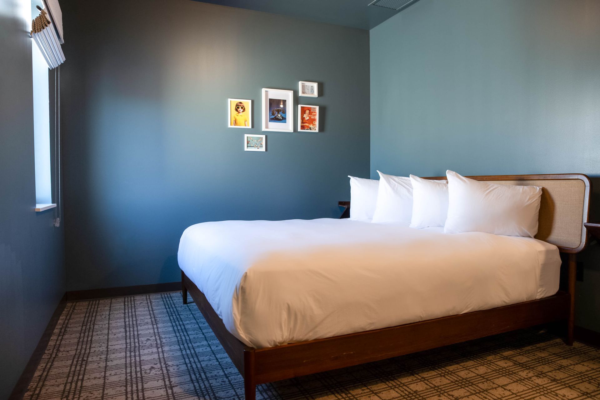 A king-sized bed with four white pillows against a teal wall in a bedroom with a small side table and lamp.