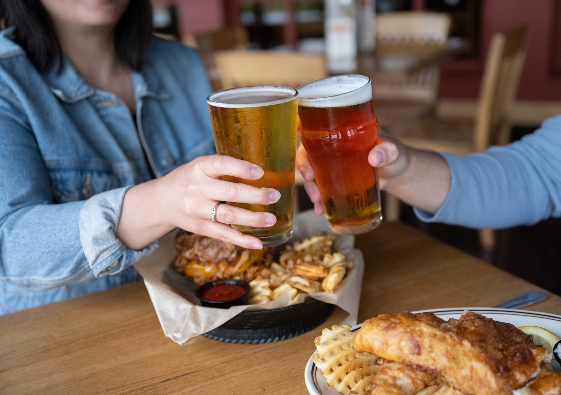 Two people clinking glasses of beer over a table filled with fish and chips and waffle fries.