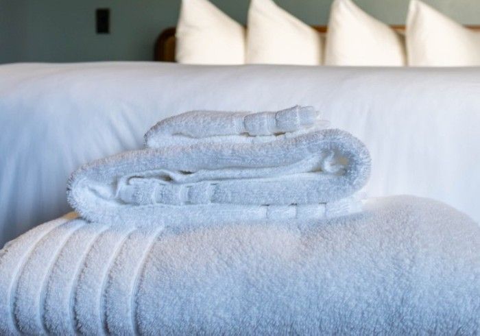 A stack of clean, white towels rests on a bed with white linens and pillows in a hotel room.