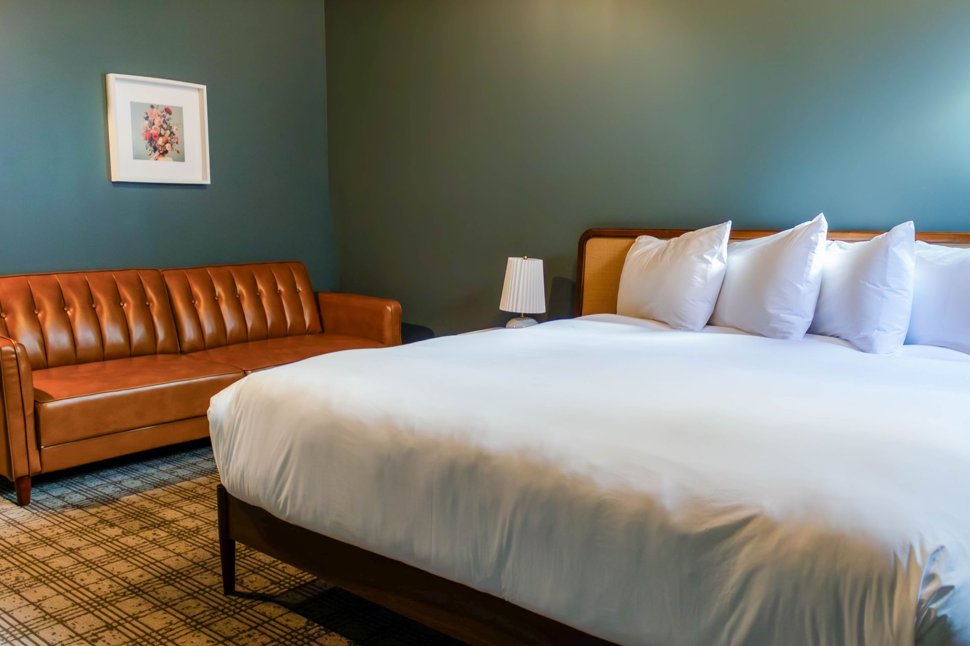 A hotel room with a large bed featuring white linens and multiple pillows next to a tan tufted leather couch.
