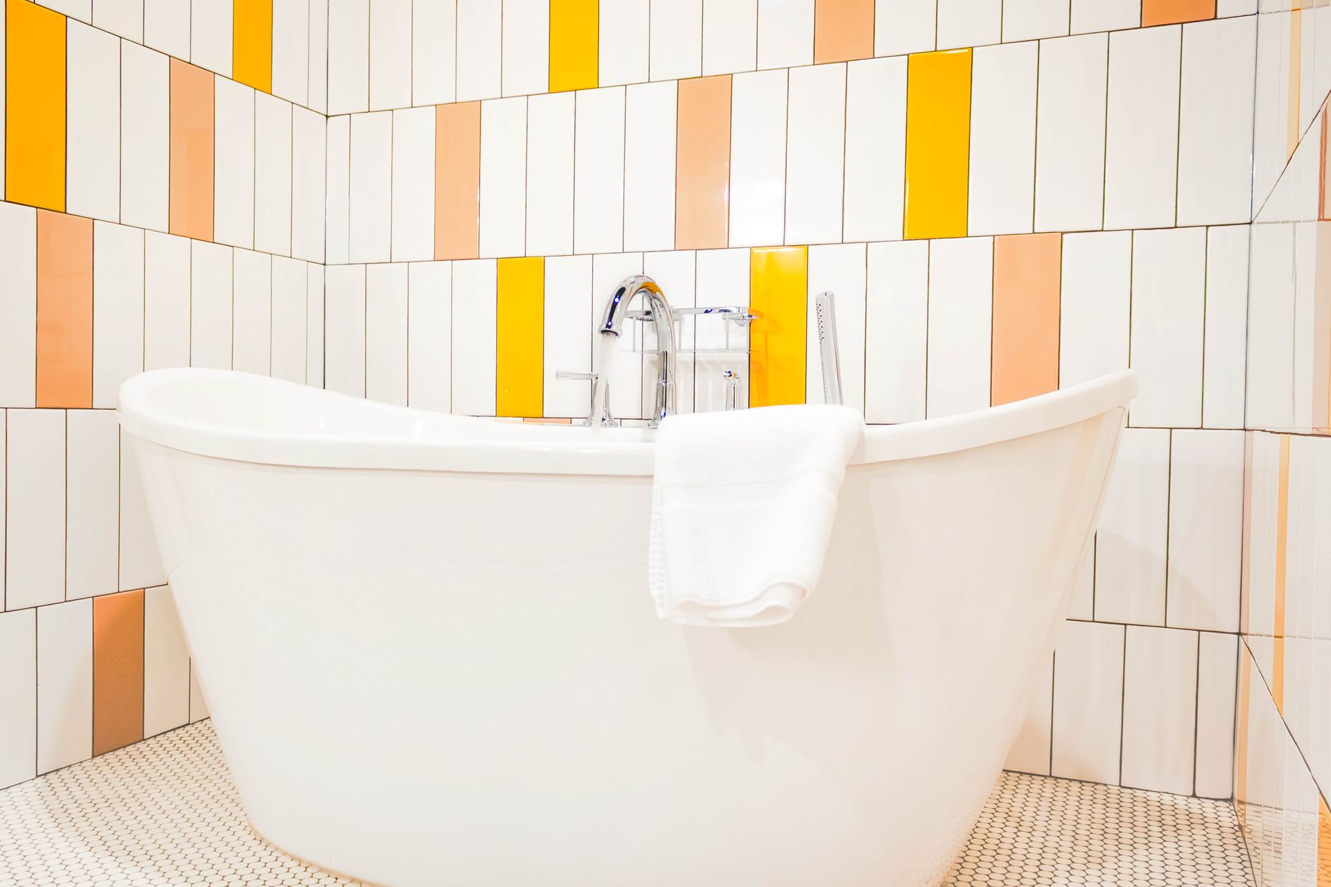 A freestanding white bathtub with a white towel, set against a tiled wall with vertical orange, yellow, and white stripes.
