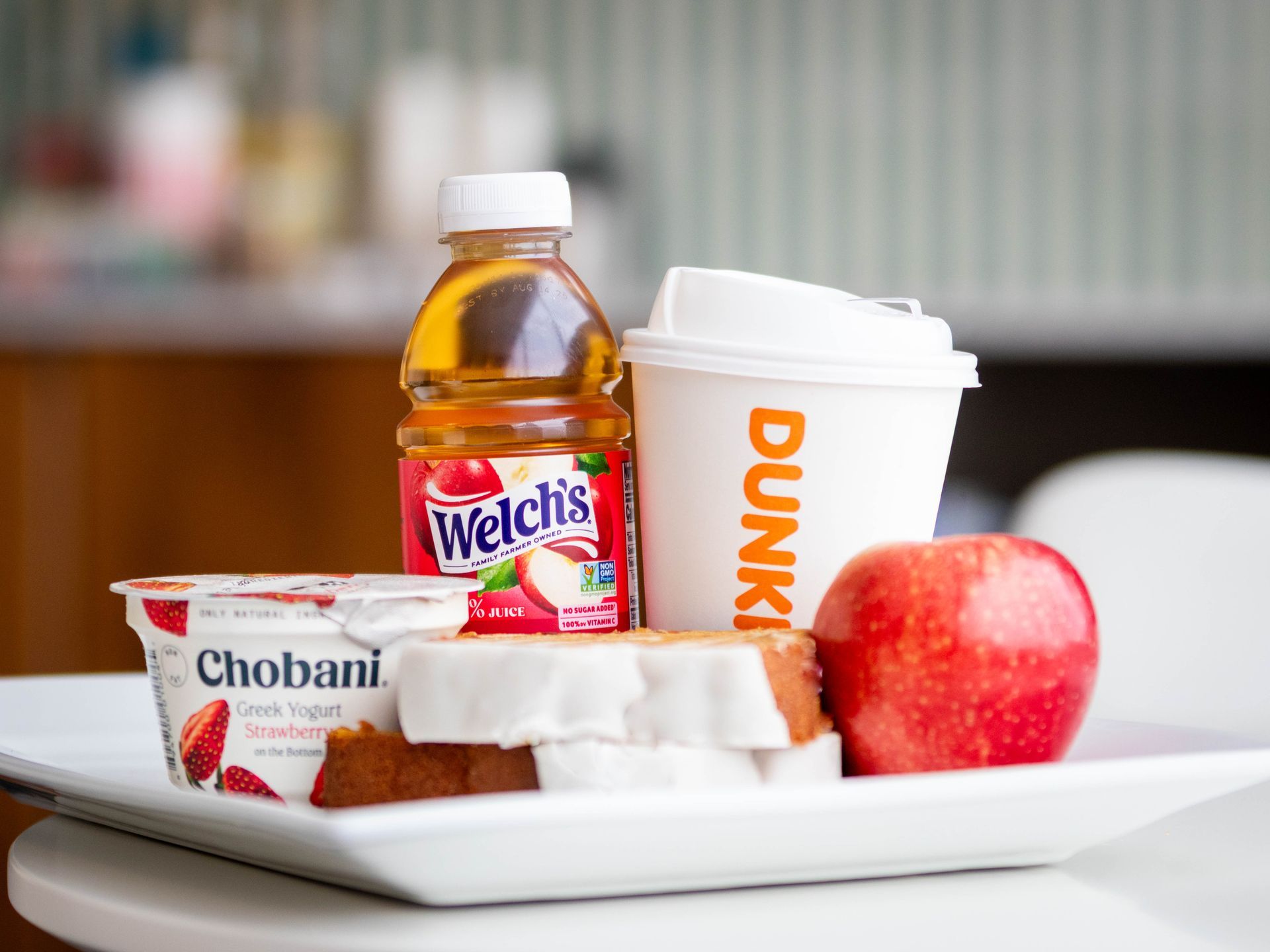 A white plate holds a Chobani yogurt, a Welch's juice bottle, a Dunkin' cup, a glazed pastry, and a red apple.