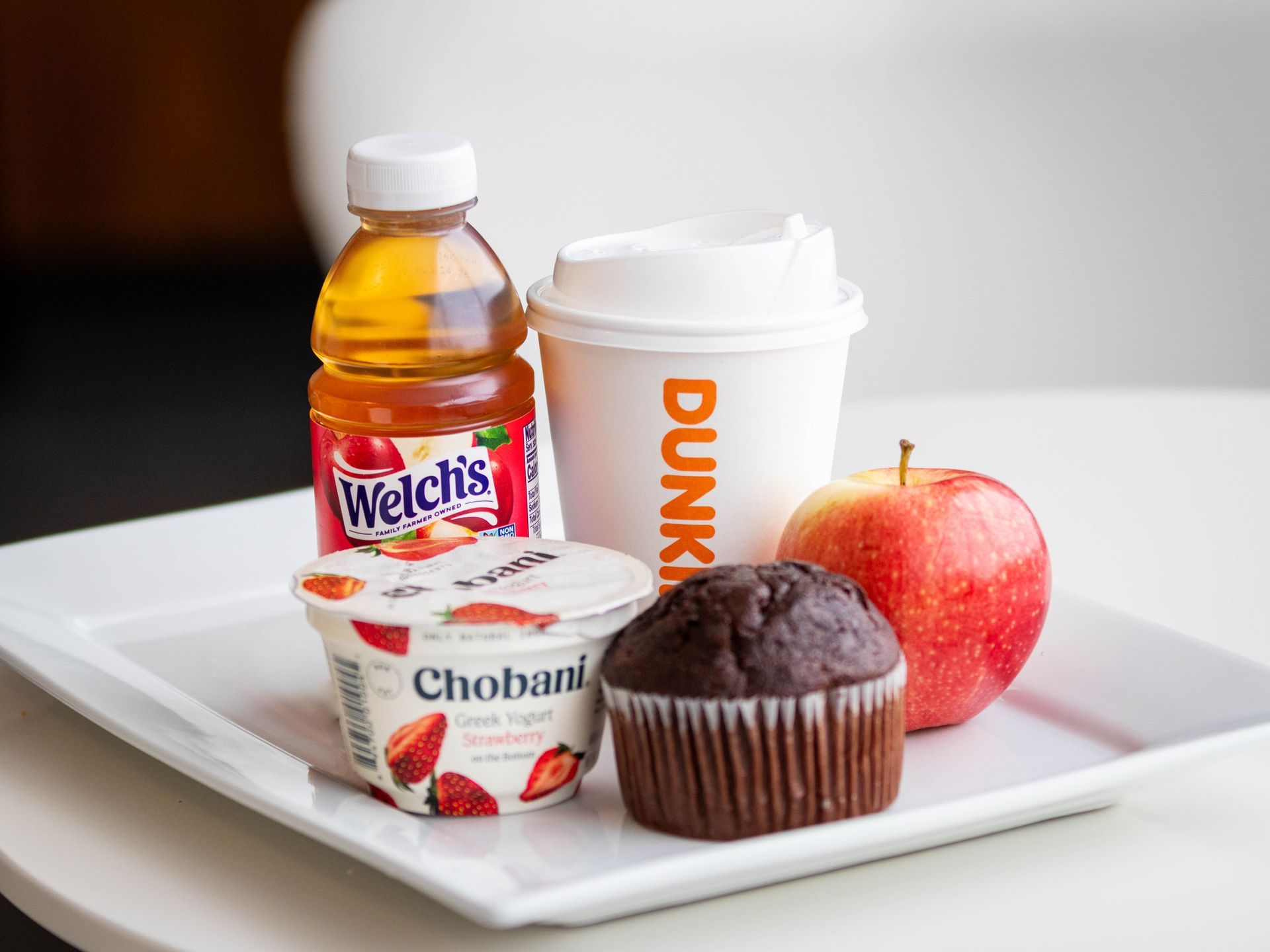 A tray holding a bottle of Welch's juice, a Dunkin' coffee cup, a strawberry Chobani yogurt, a muffin, and an apple.