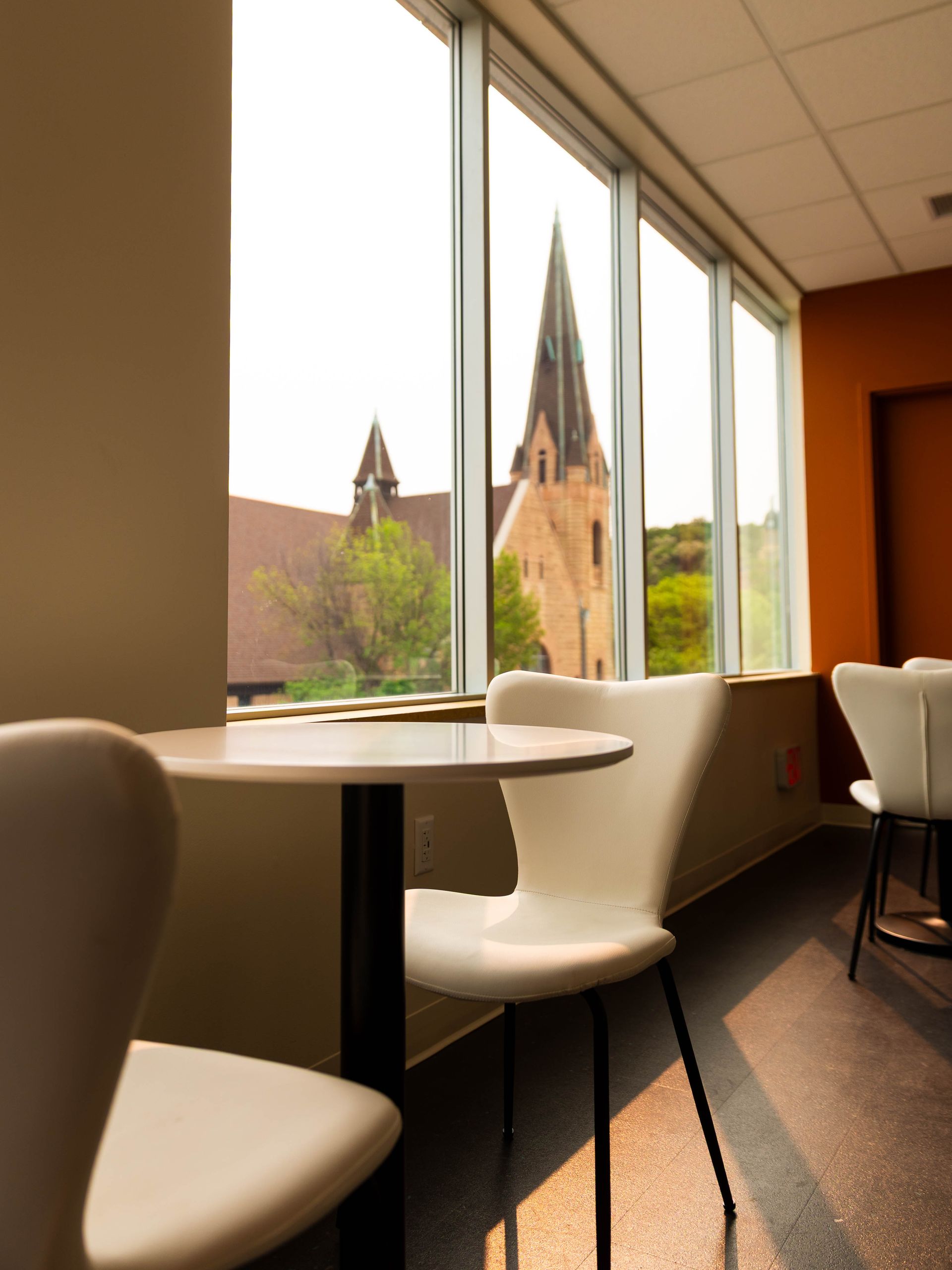 A round table and white chairs in a room with large windows overlooking a historic stone church with a tall steeple.