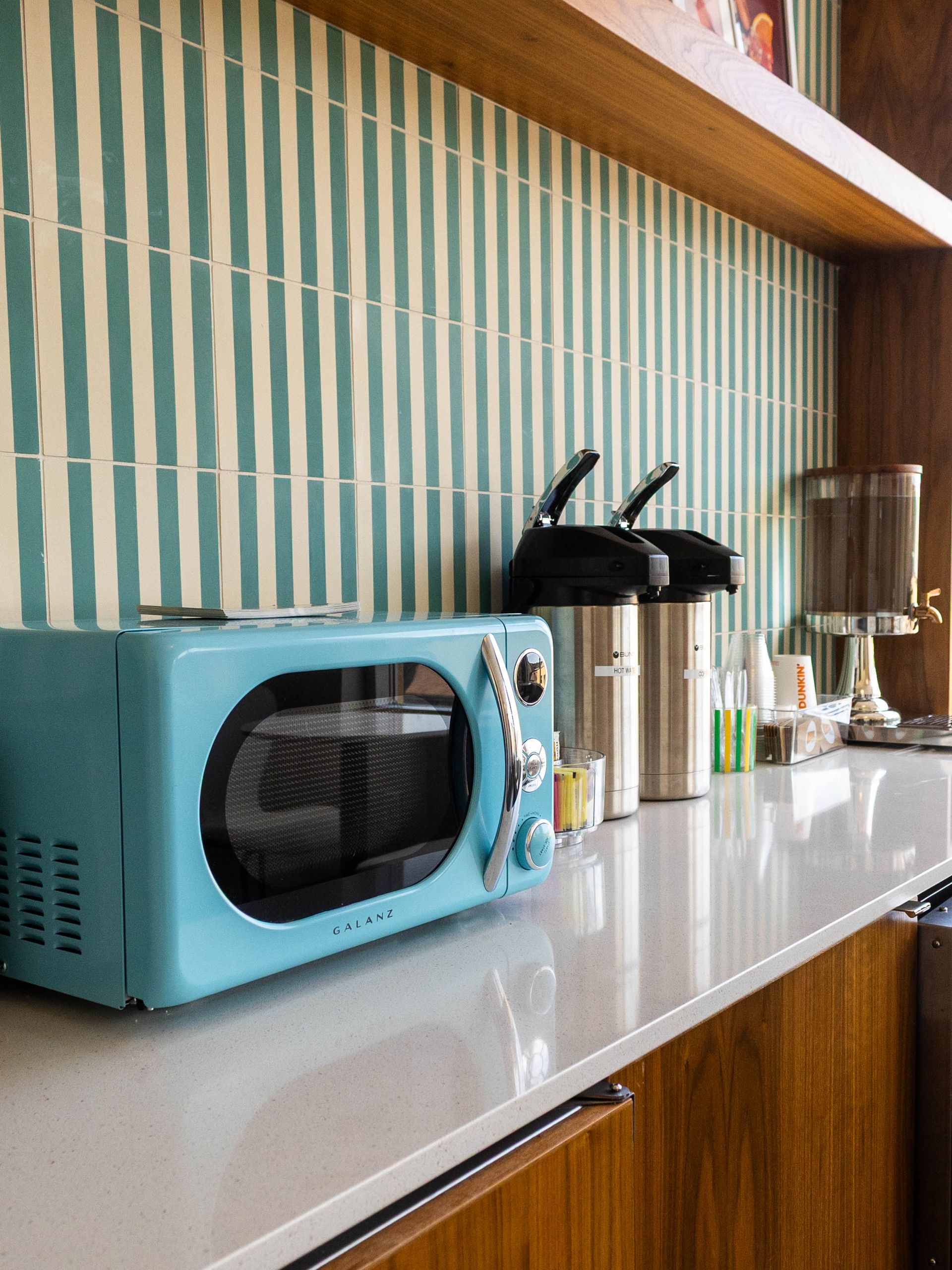 A turquoise retro microwave sits on a white countertop next to two beverage dispensers against a teal striped tile wall.