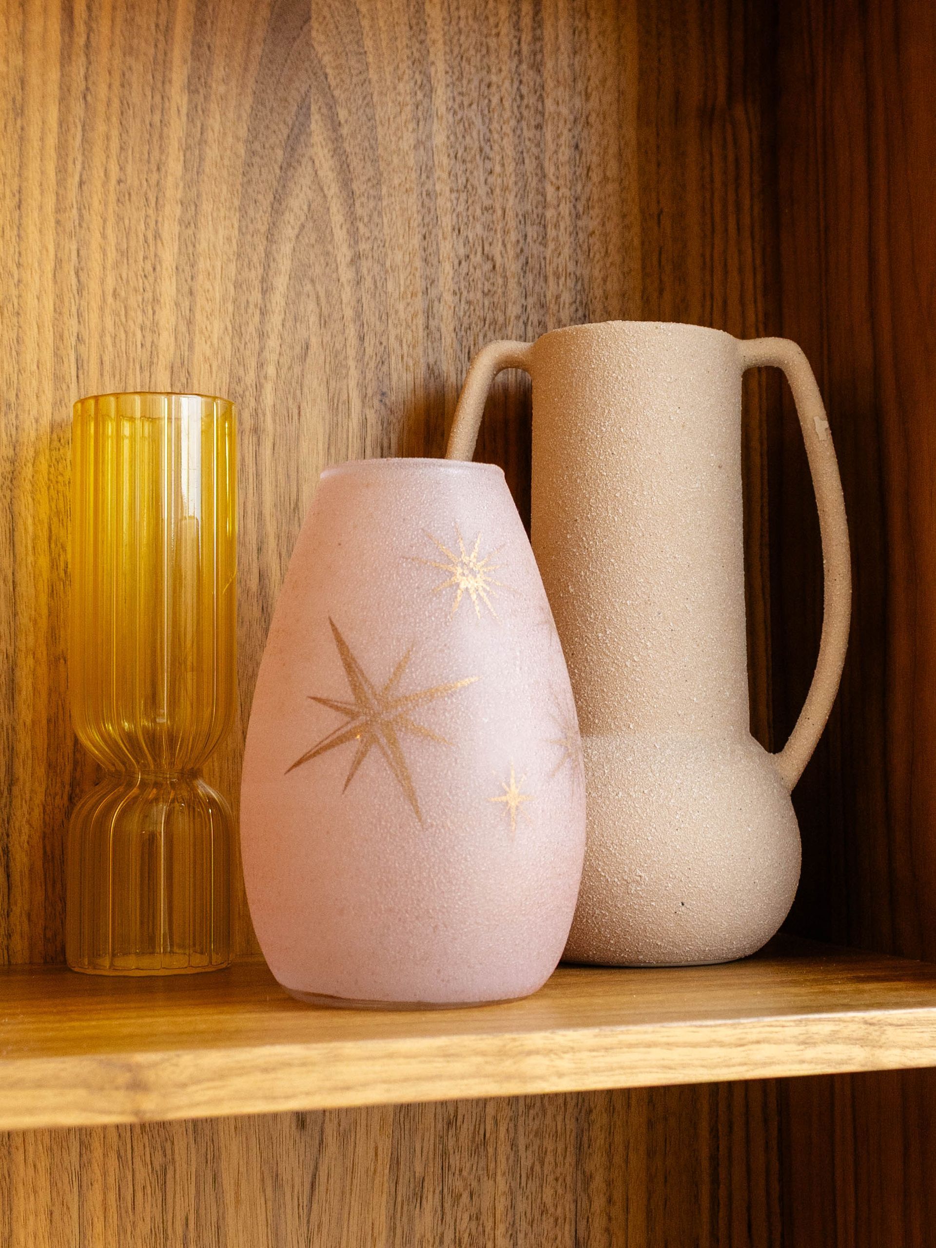 Three decorative vases—one yellow glass, one frosted pink with starburst patterns, and one textured beige—on a wood shelf.