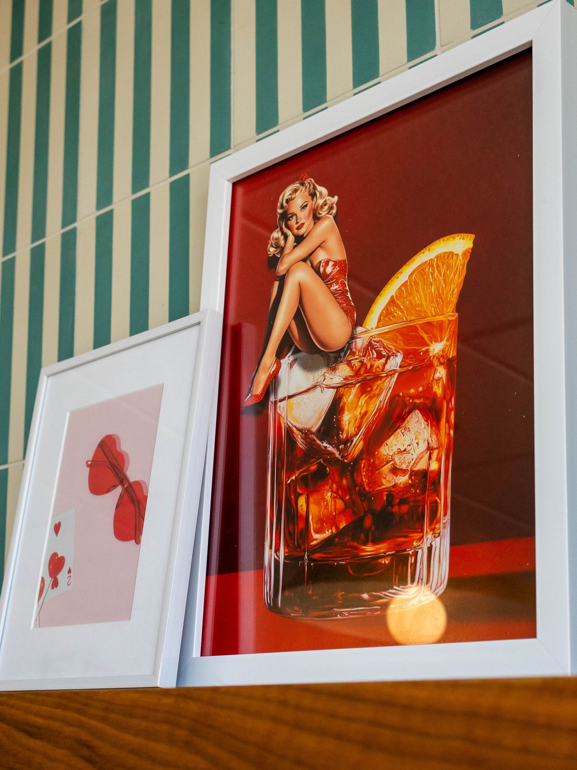 Two framed art prints on a shelf: one featuring a pin-up style illustration in a cocktail, the other with a red bow.