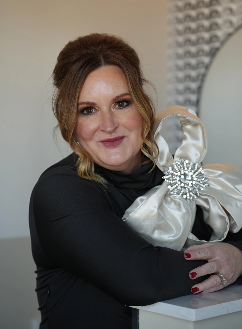 A person with auburn hair in an updo wears a black top and a large, white fabric bow accessory with a crystal center.