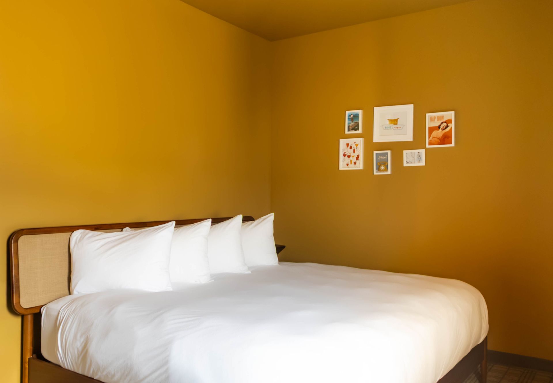 A made bed with white pillows and a duvet against a mustard-yellow wall with a small gallery of framed art.
