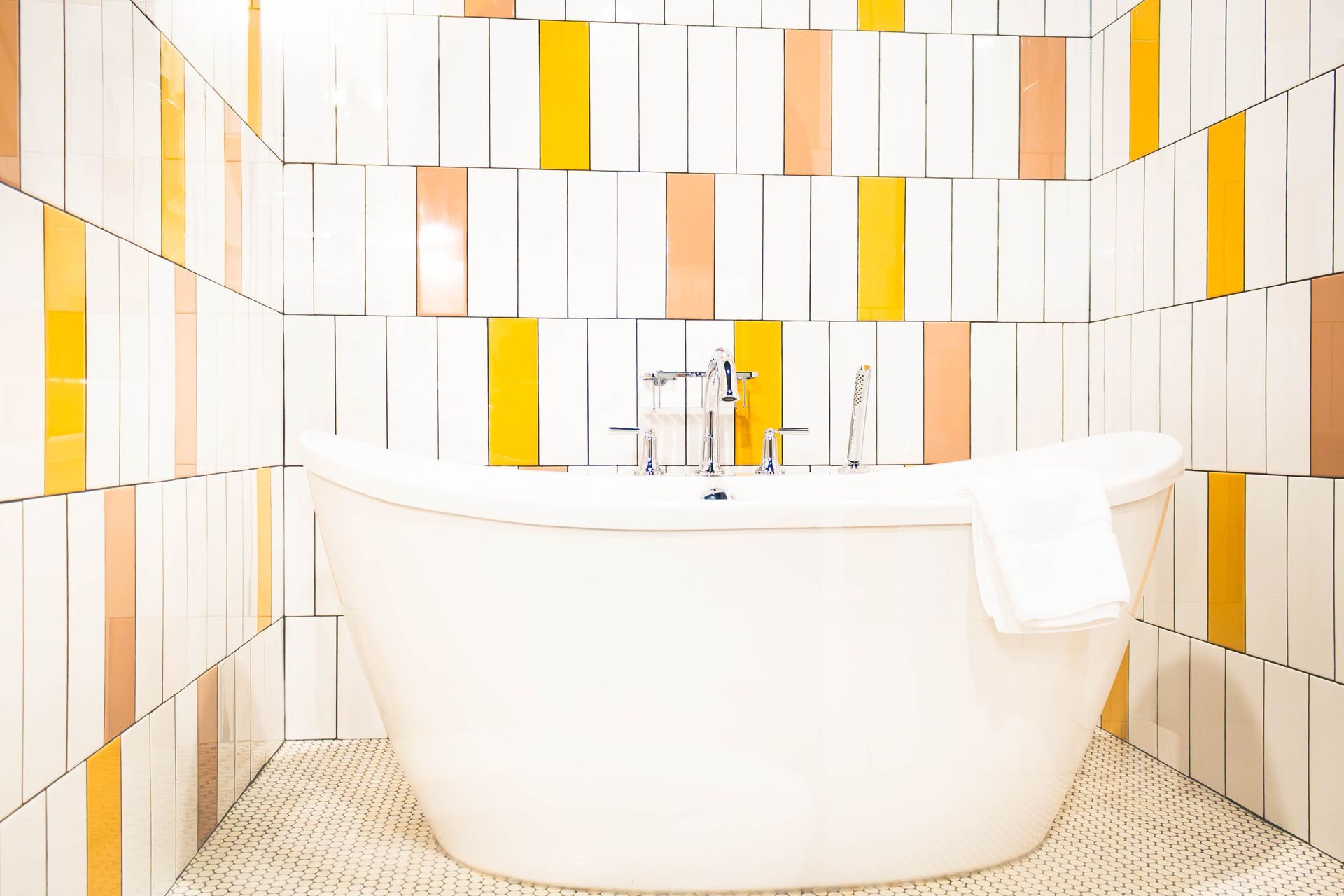 A white freestanding bathtub sits against a wall of vertical tiles in shades of white, yellow, and peach.