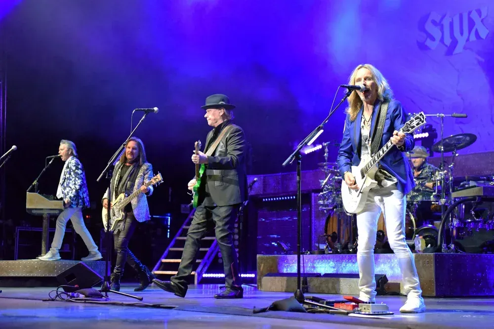 The band Styx performs on a concert stage with blue lighting; musicians play guitars and keyboards under spotlights.